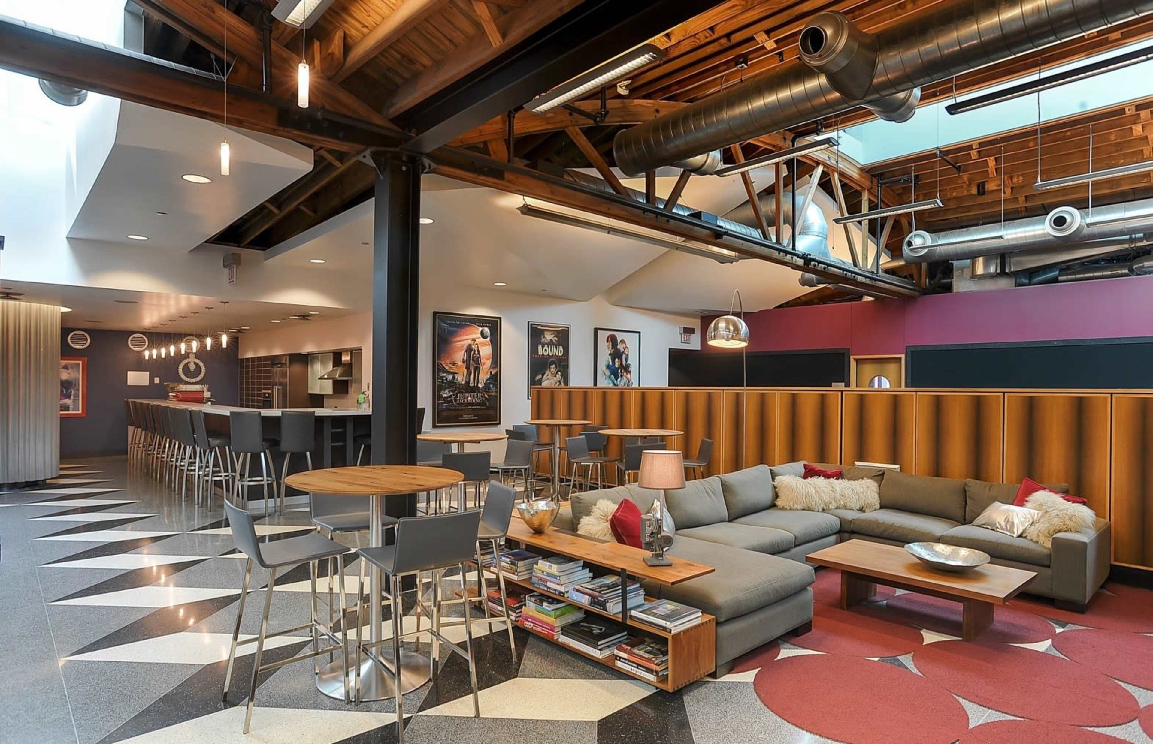 A modern lounge area featuring a mix of seating options, a bar with high stools, and decorative wall posters in a spacious interior with wooden beams and exposed ducts.