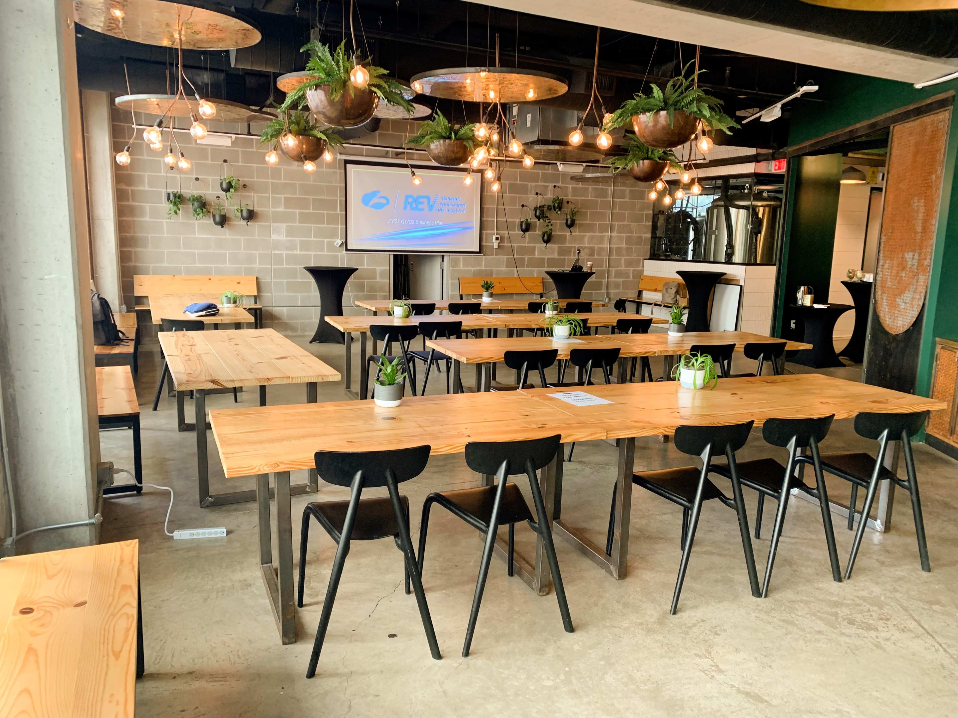 The image shows a modern conference room arranged with long wooden tables and chairs, featuring plants and a projector screen.