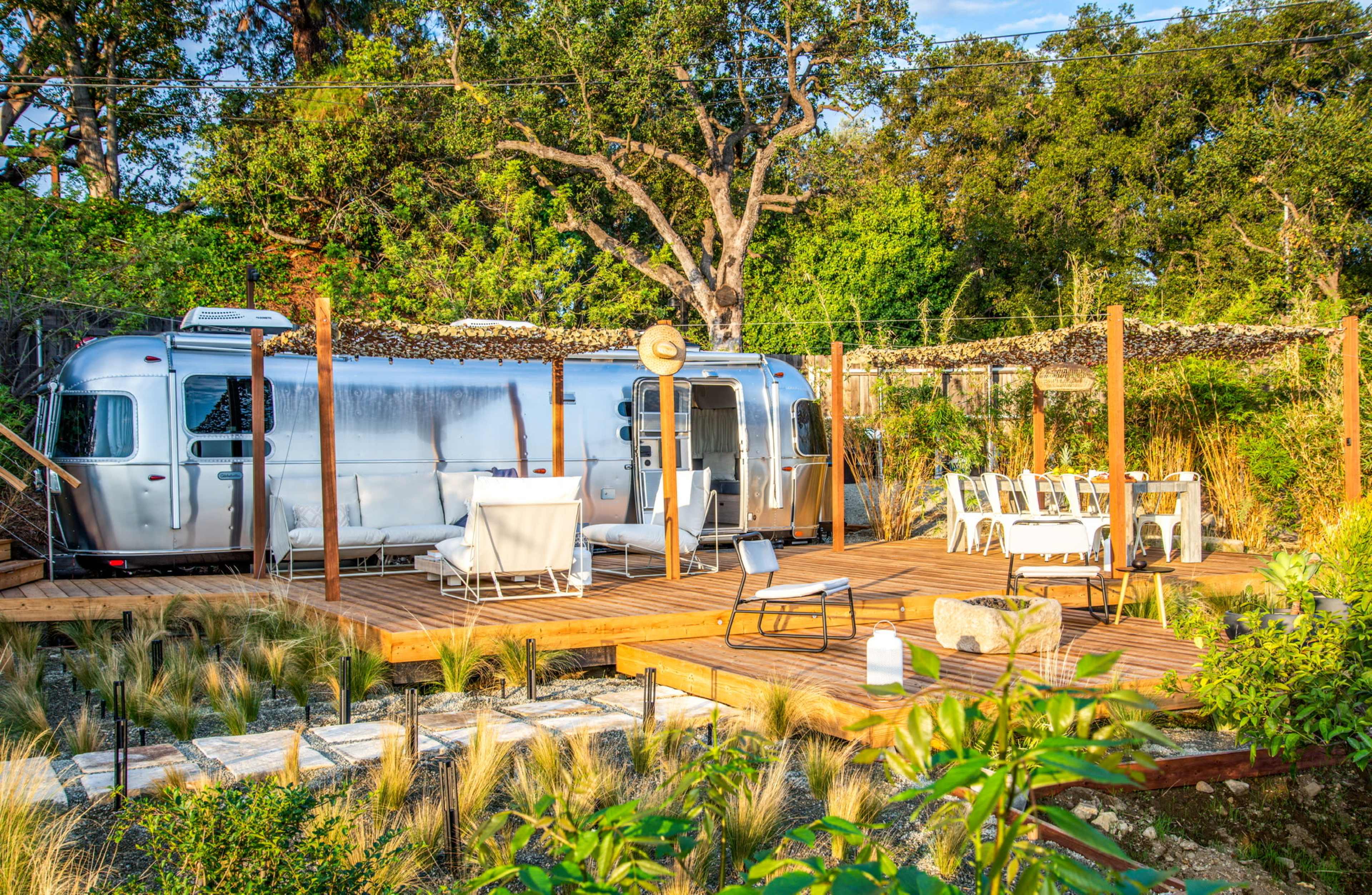 A silver Airstream trailer is parked on a wooden deck surrounded by scattered seating and dining furniture in a landscaped backyard.