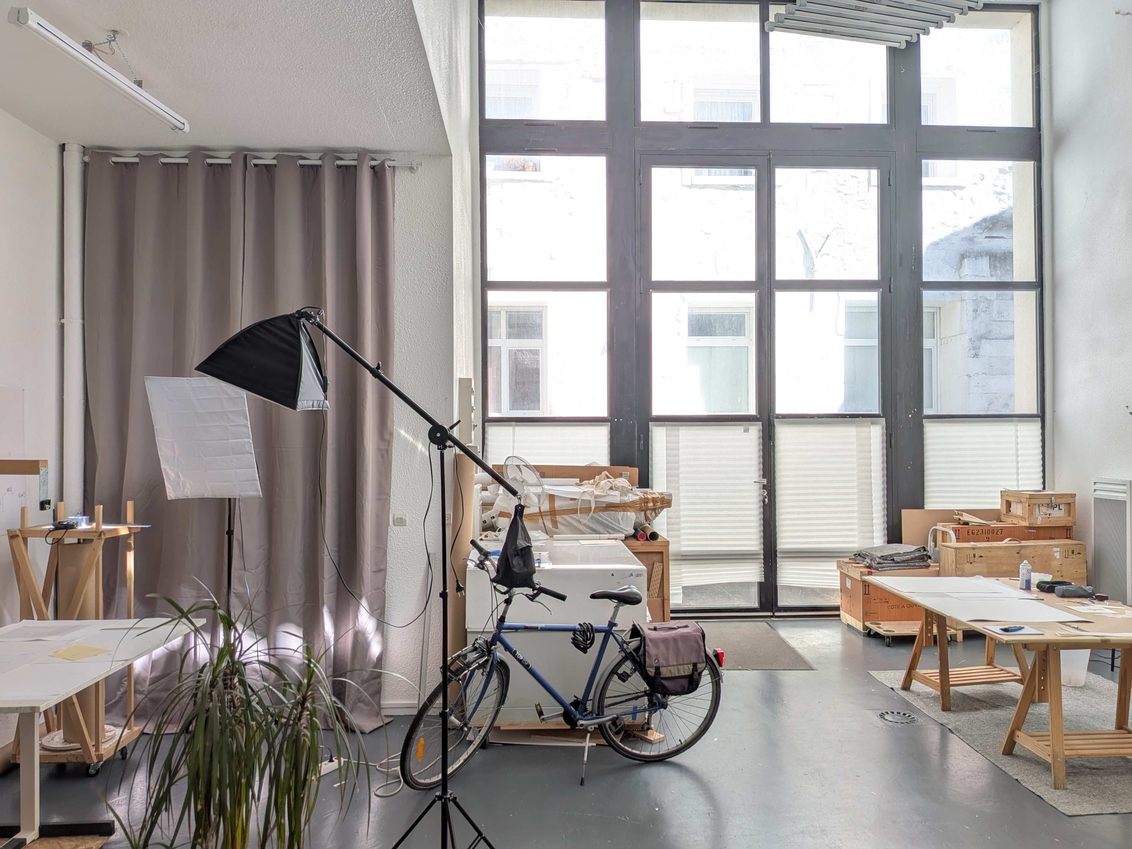The image shows a bright studio space with large windows, a bicycle, a photography light, and wooden furniture arranged around the room.