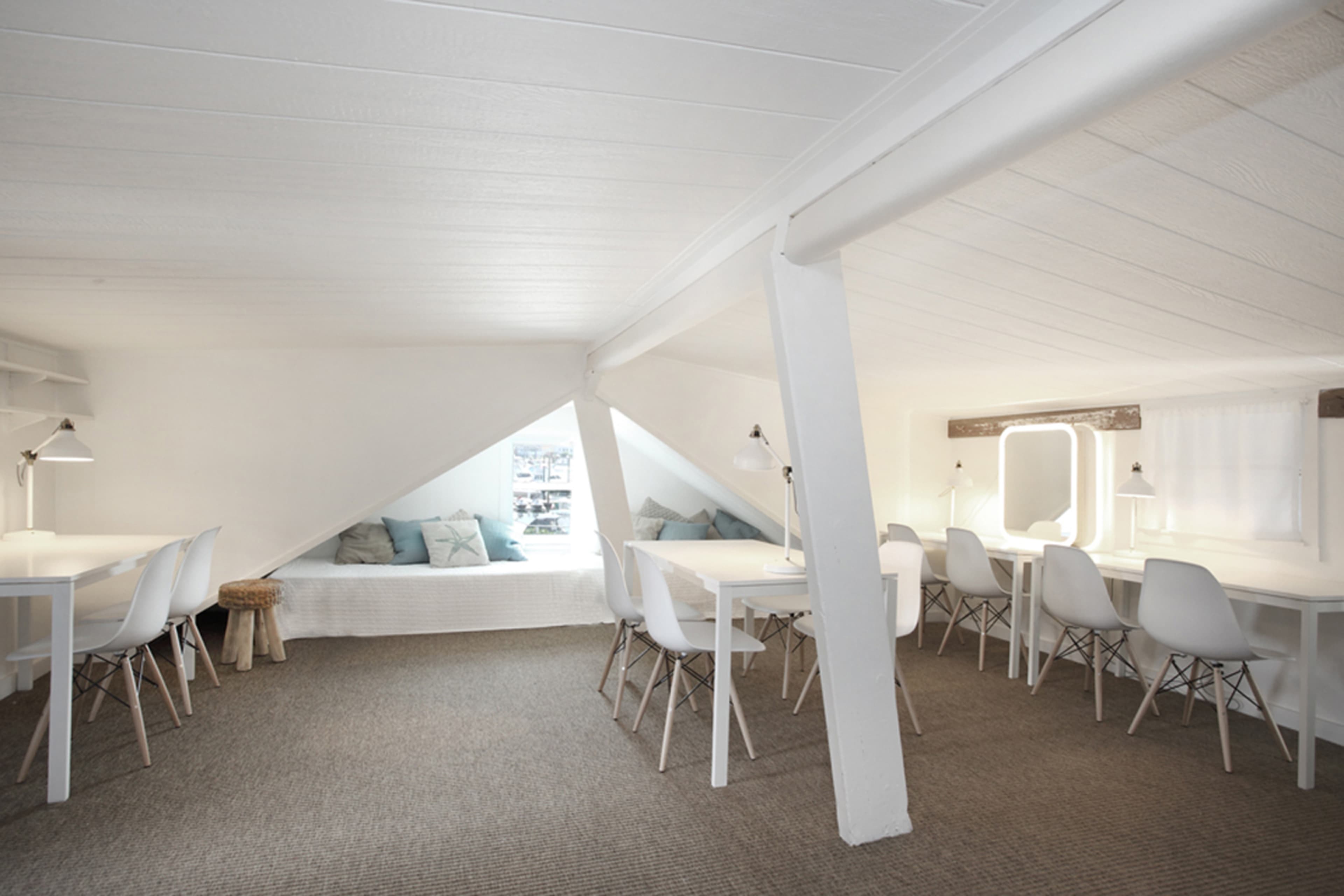A bright, minimalist attic space features white furniture, a corner seating area with cushions, and large windows that allow natural light to illuminate the room.