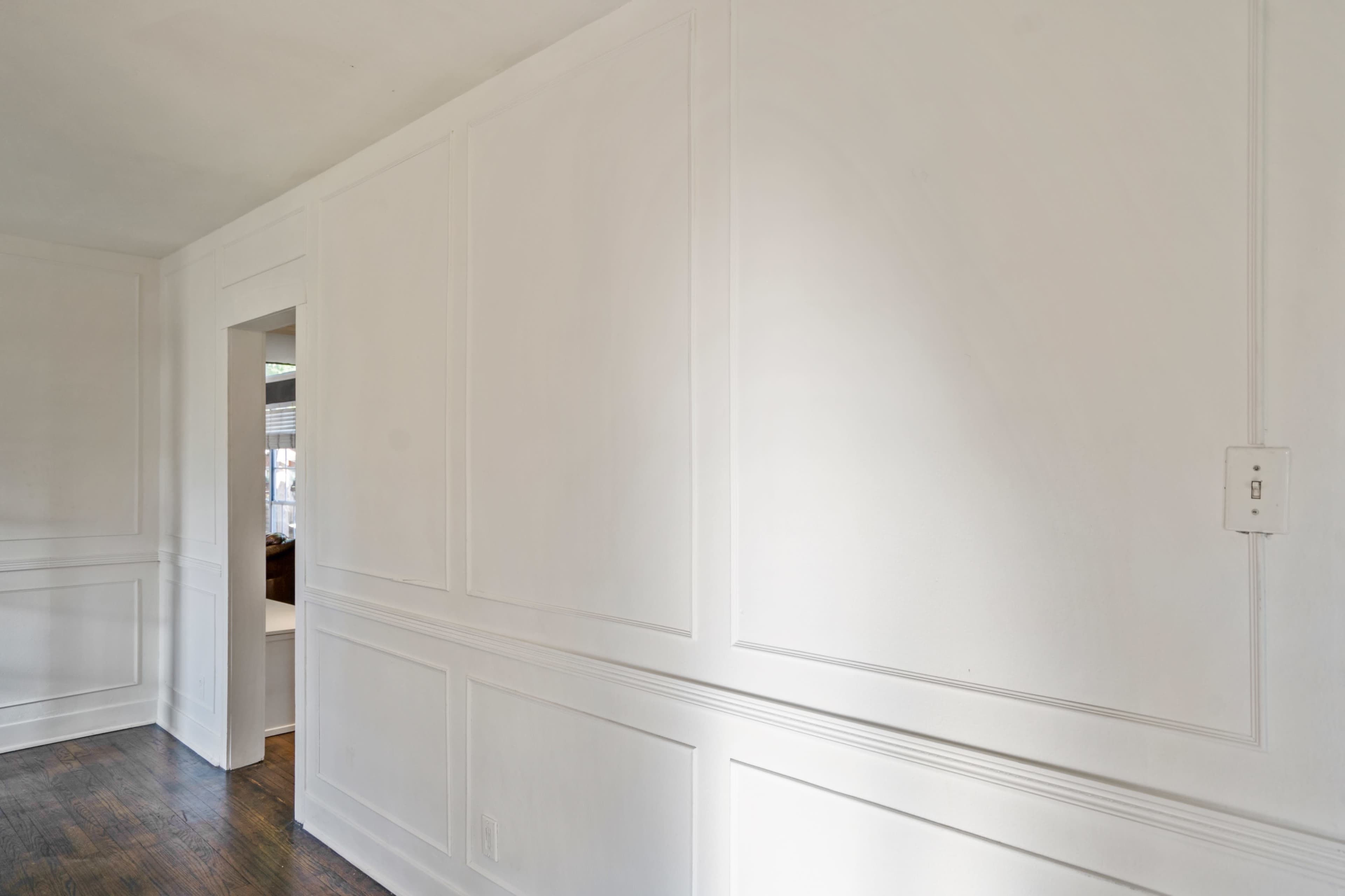 A bright, white wall with paneling and a doorway leading to another room.