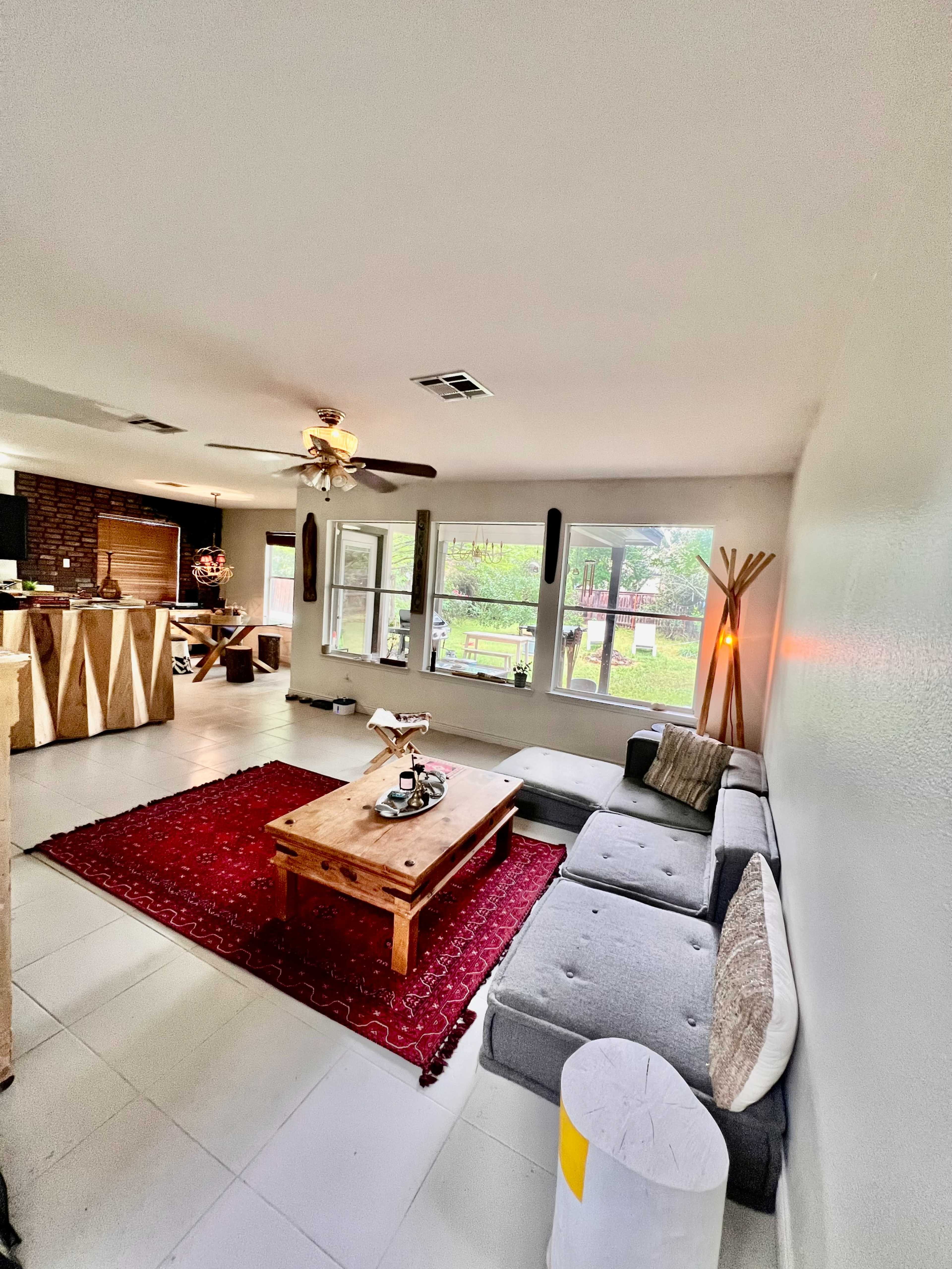 A living room with a gray sectional sofa, a wooden coffee table on a red area rug, and large windows providing natural light.