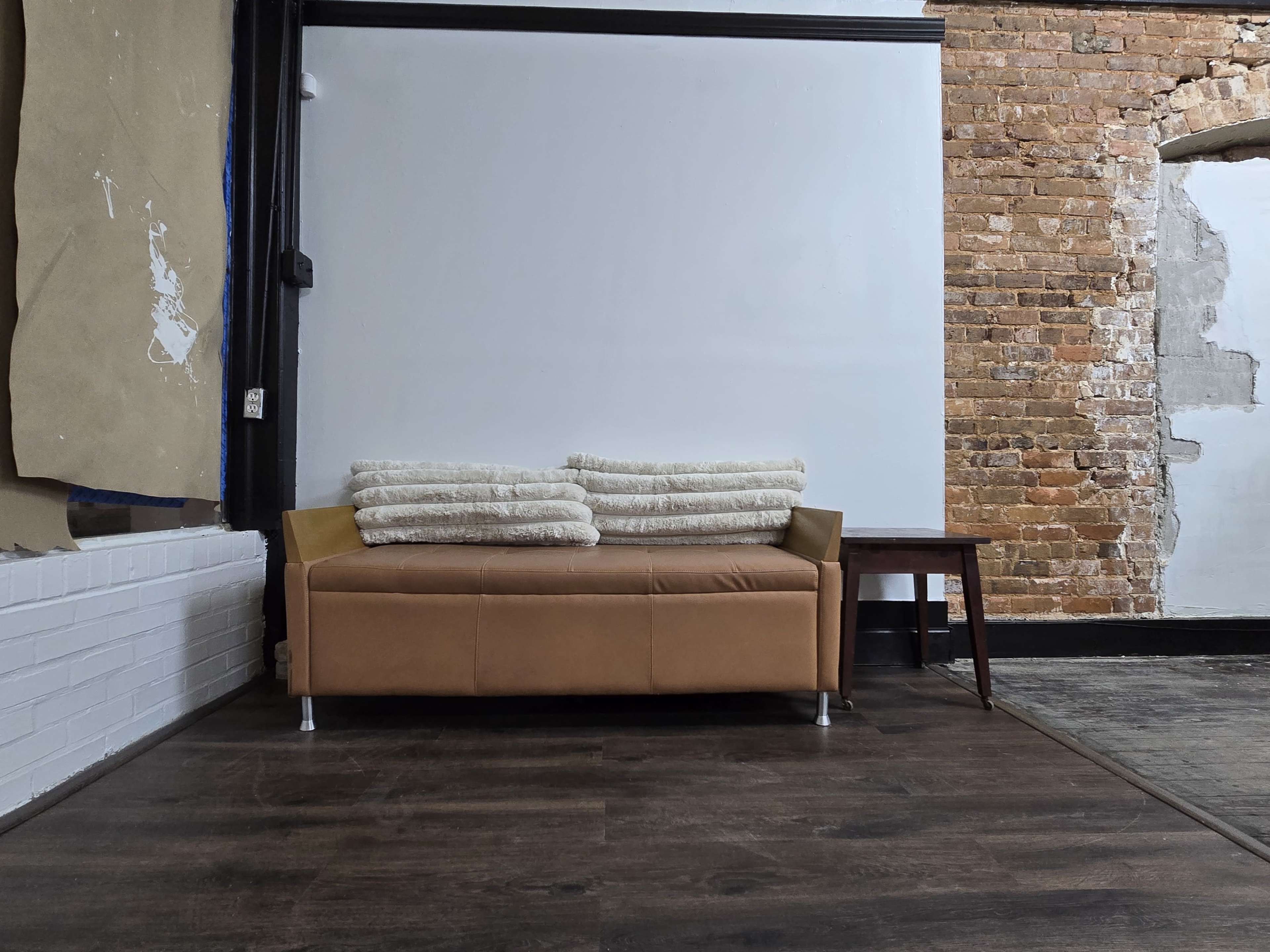 A brown leather sofa with rolled blankets rests against a white wall in a space with exposed brick and wooden flooring.