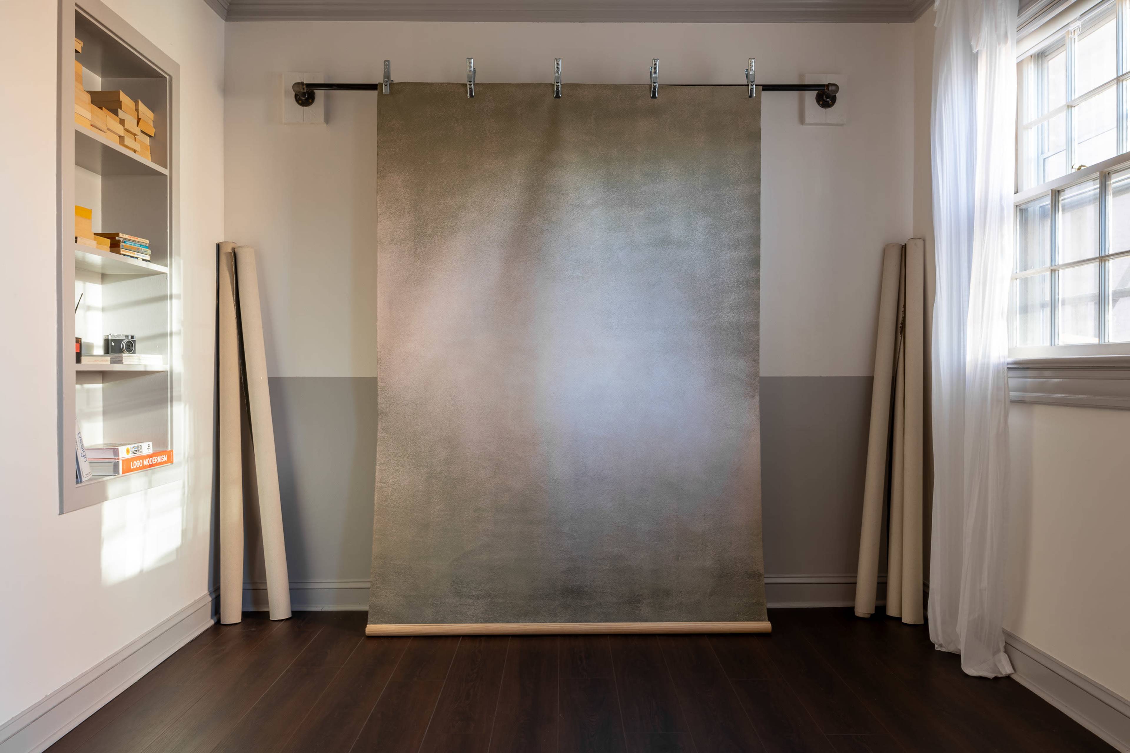 A plain backdrop is hung on a wall in a well-lit room with wooden flooring and shelves filled with various items.