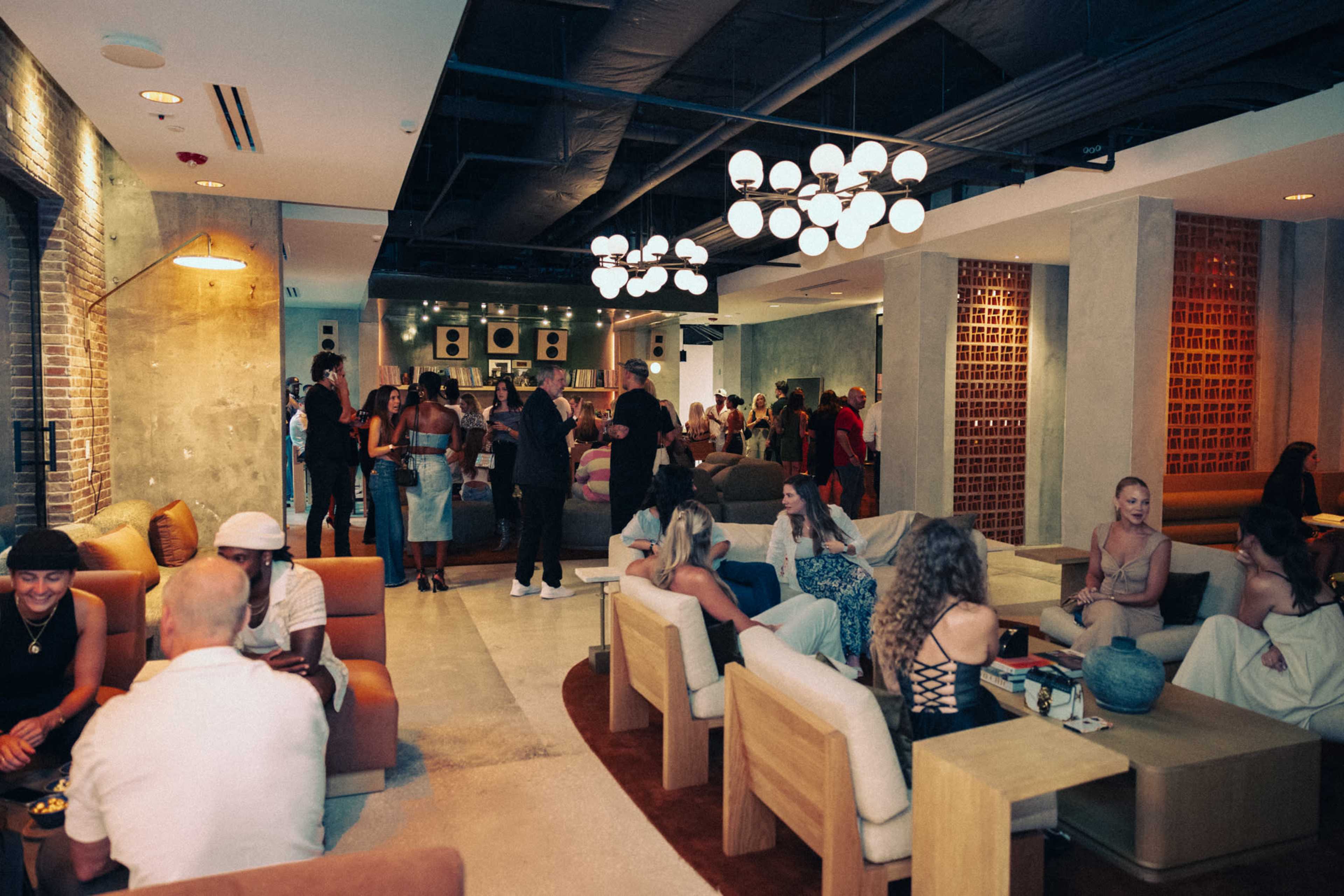 The image shows a lively lounge with groups of people socializing, seated on modern furniture surrounded by industrial-style decor.