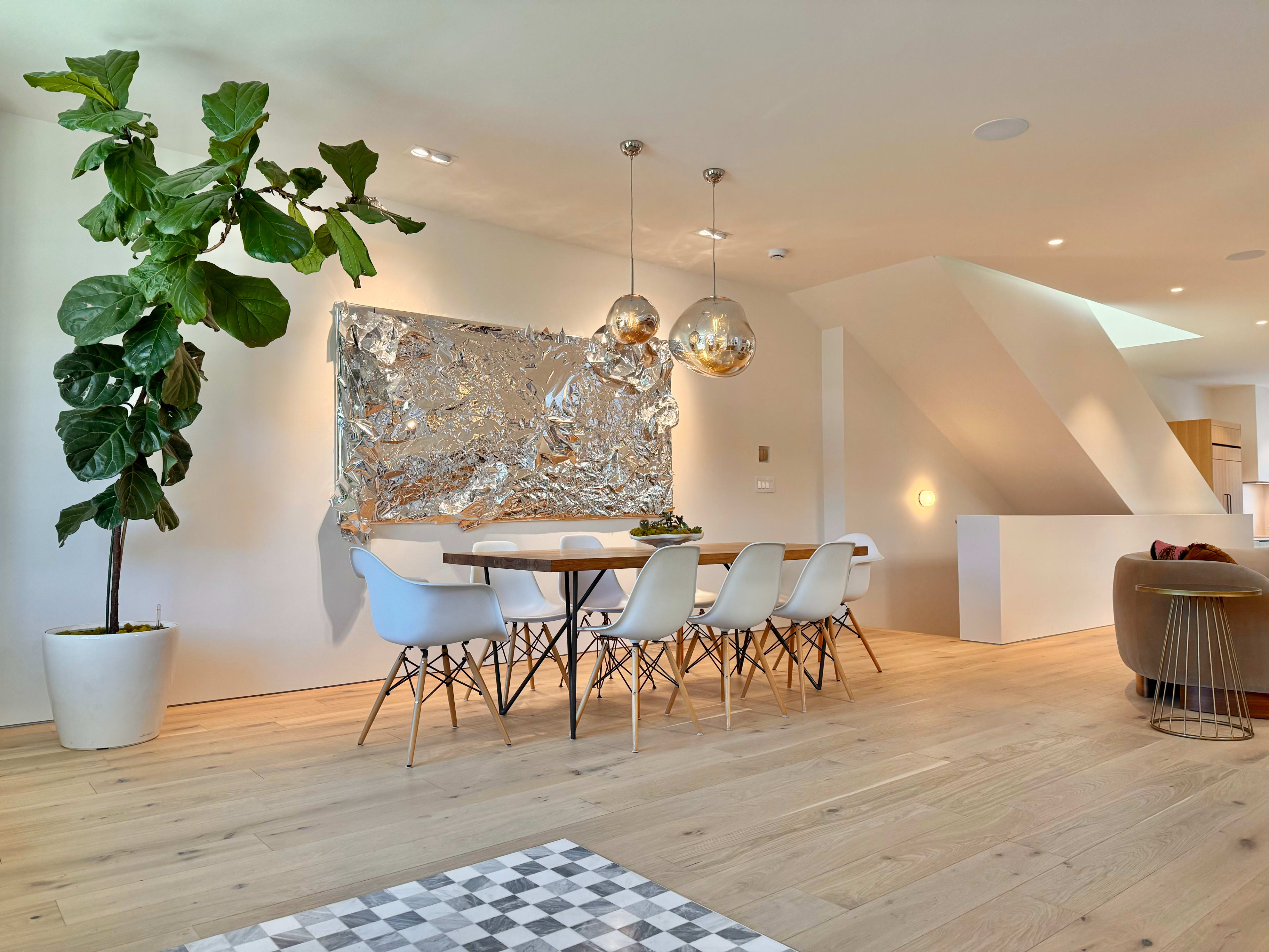The image shows a modern dining area with a long table surrounded by white chairs, a large piece of artwork on the wall, and two pendant lights hanging above the table.