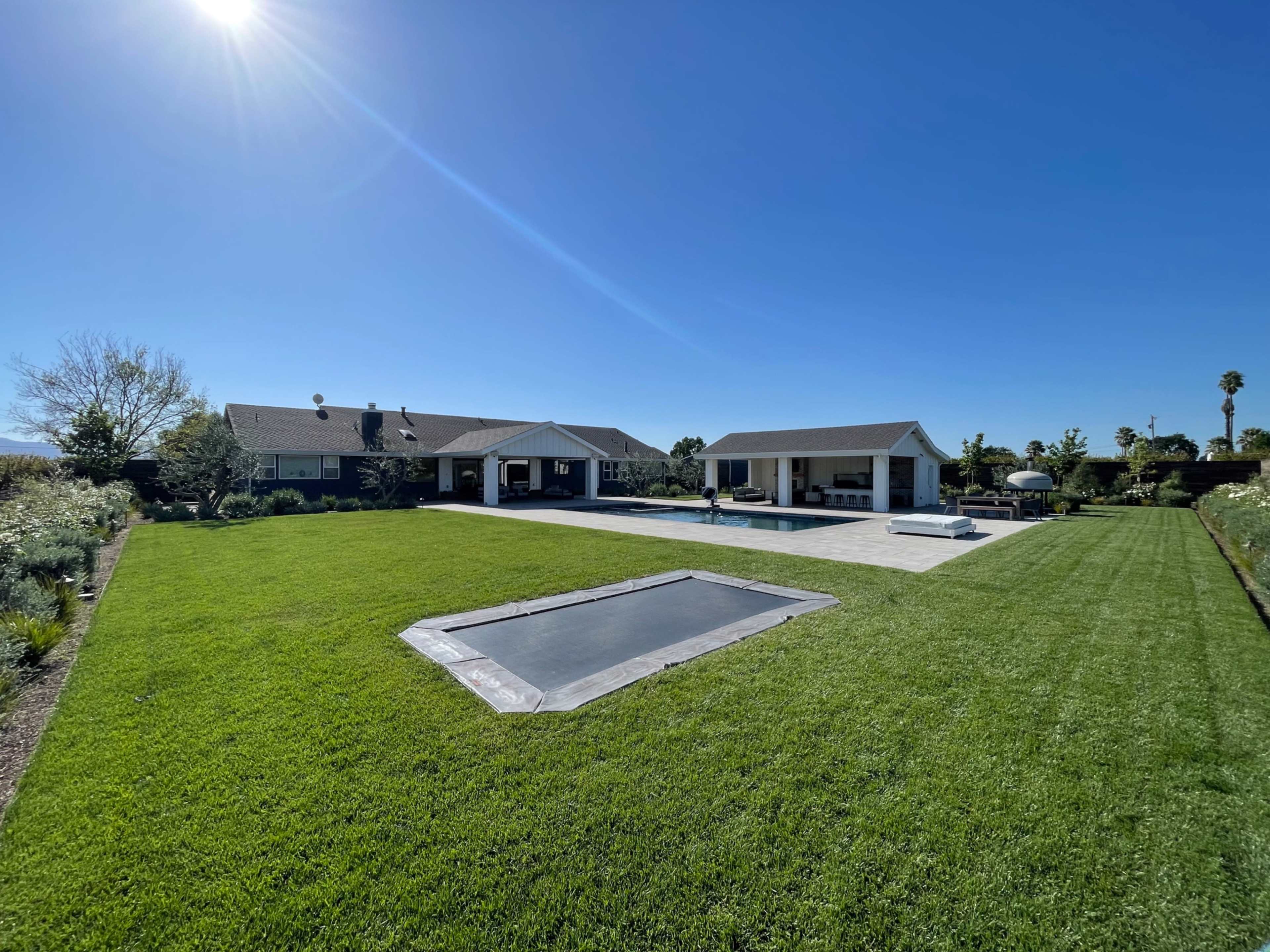 A spacious backyard features a green lawn, a swimming pool covered with a tarp, and two adjacent buildings under a clear blue sky.