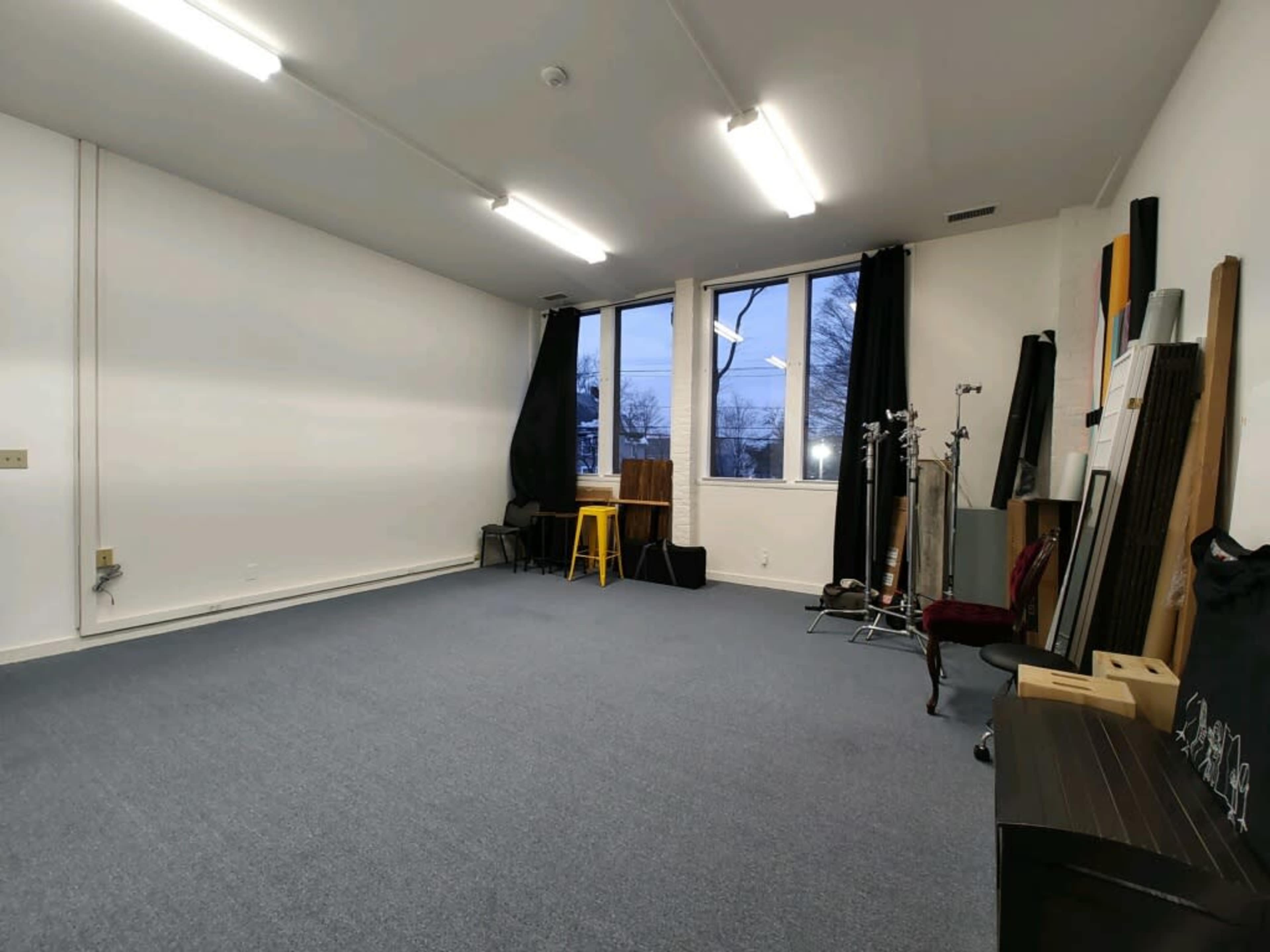 A spacious, empty room with blue carpeting, two large windows, and various equipment and furniture positioned along the walls.