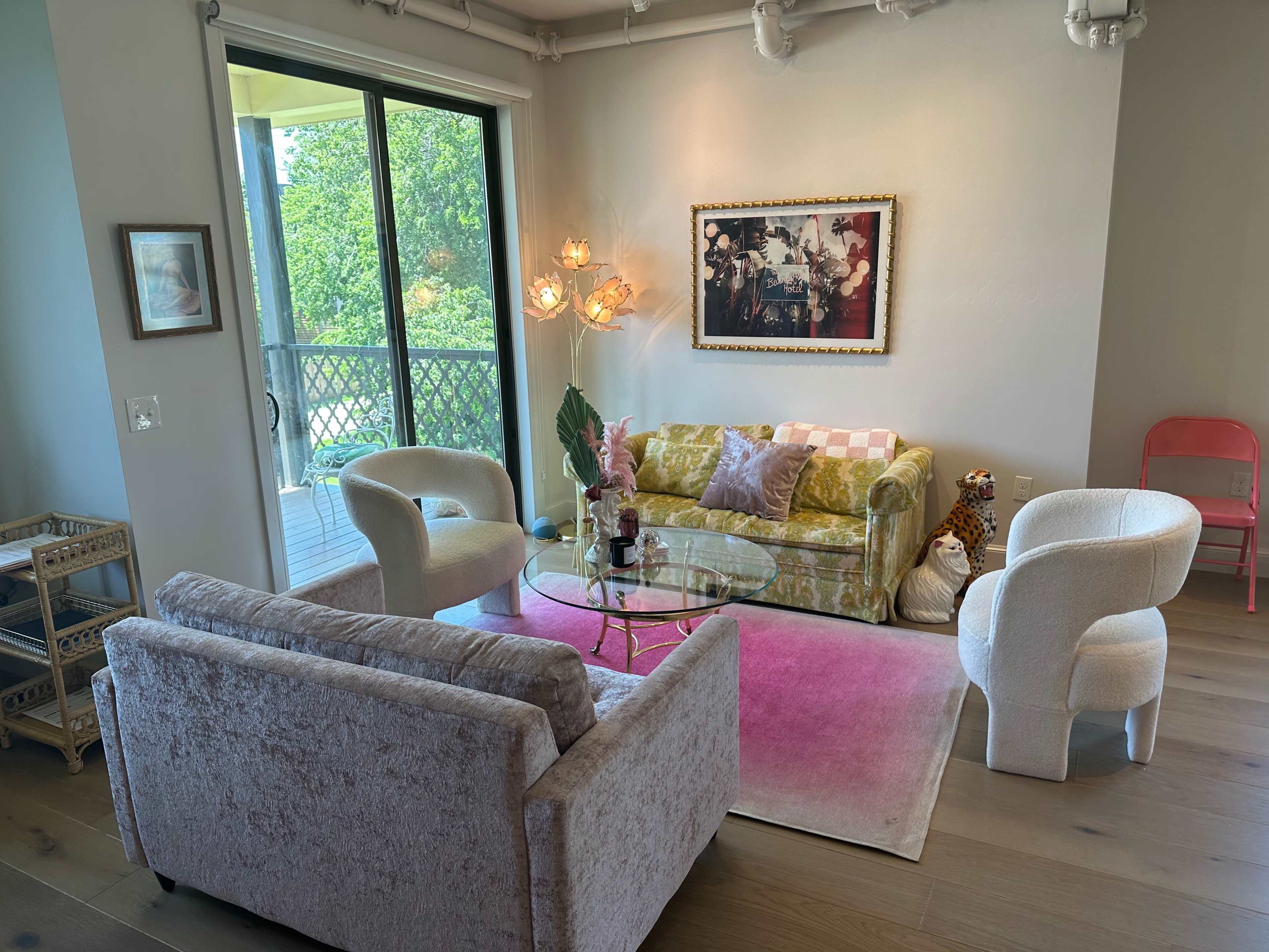 A living room with a green and yellow sofa, two white armchairs, a circular glass coffee table, and a pink rug, all illuminated by natural light from large windows.