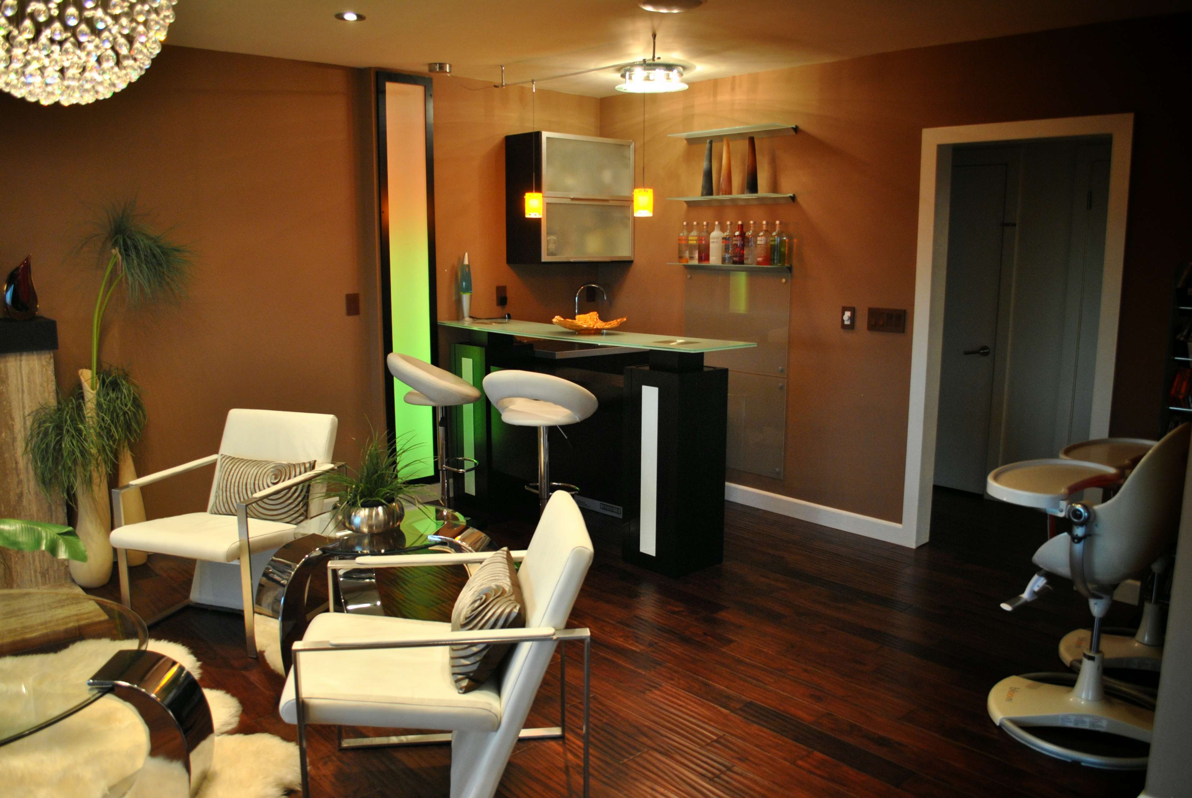 A modern living space featuring a sleek bar area with stools, white armchairs, and a glass table, complemented by wood flooring and a crystal chandelier.