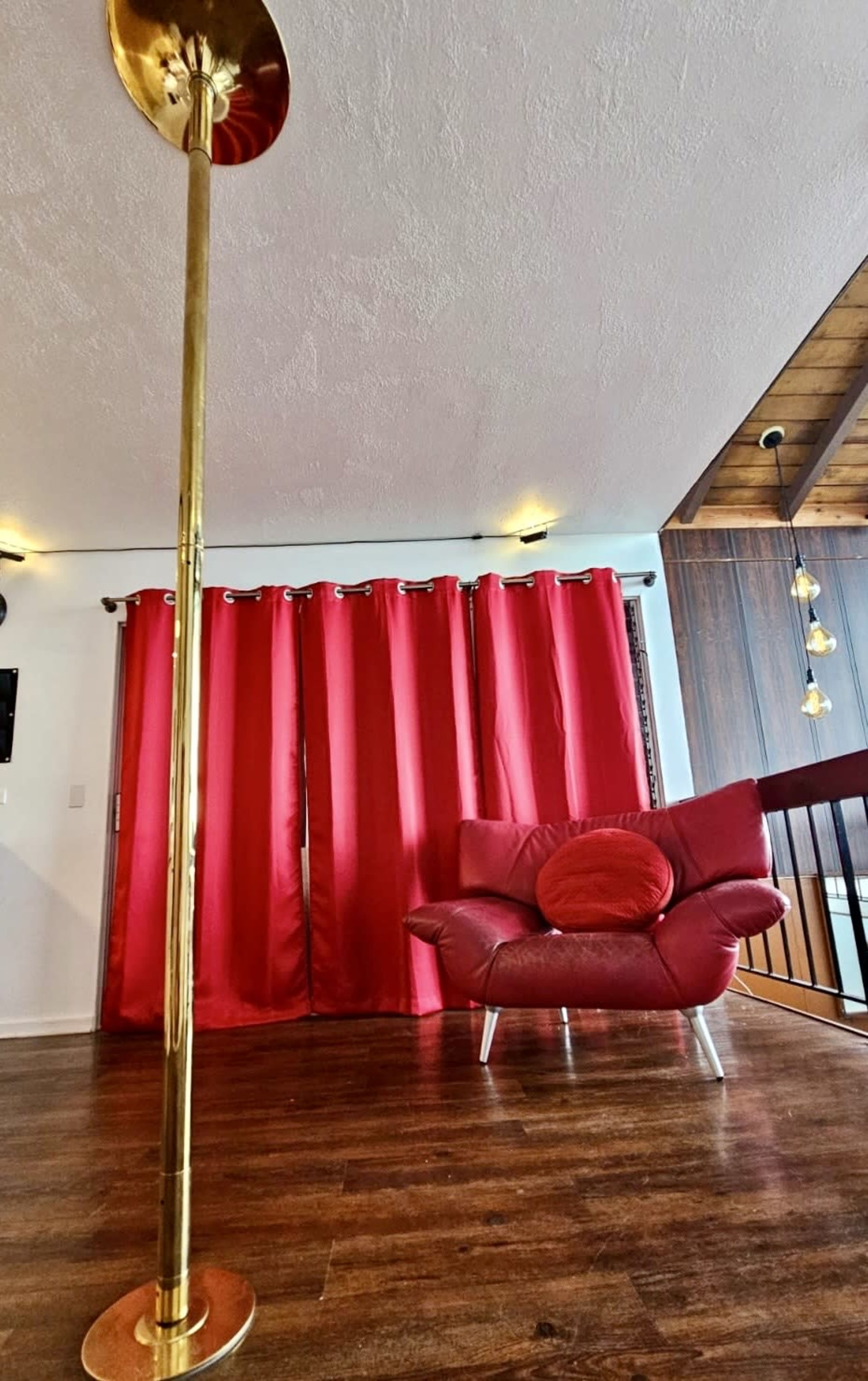 A brass floor lamp stands beside a red curtain, overlooking a modern chair with a round cushion in a living room space.