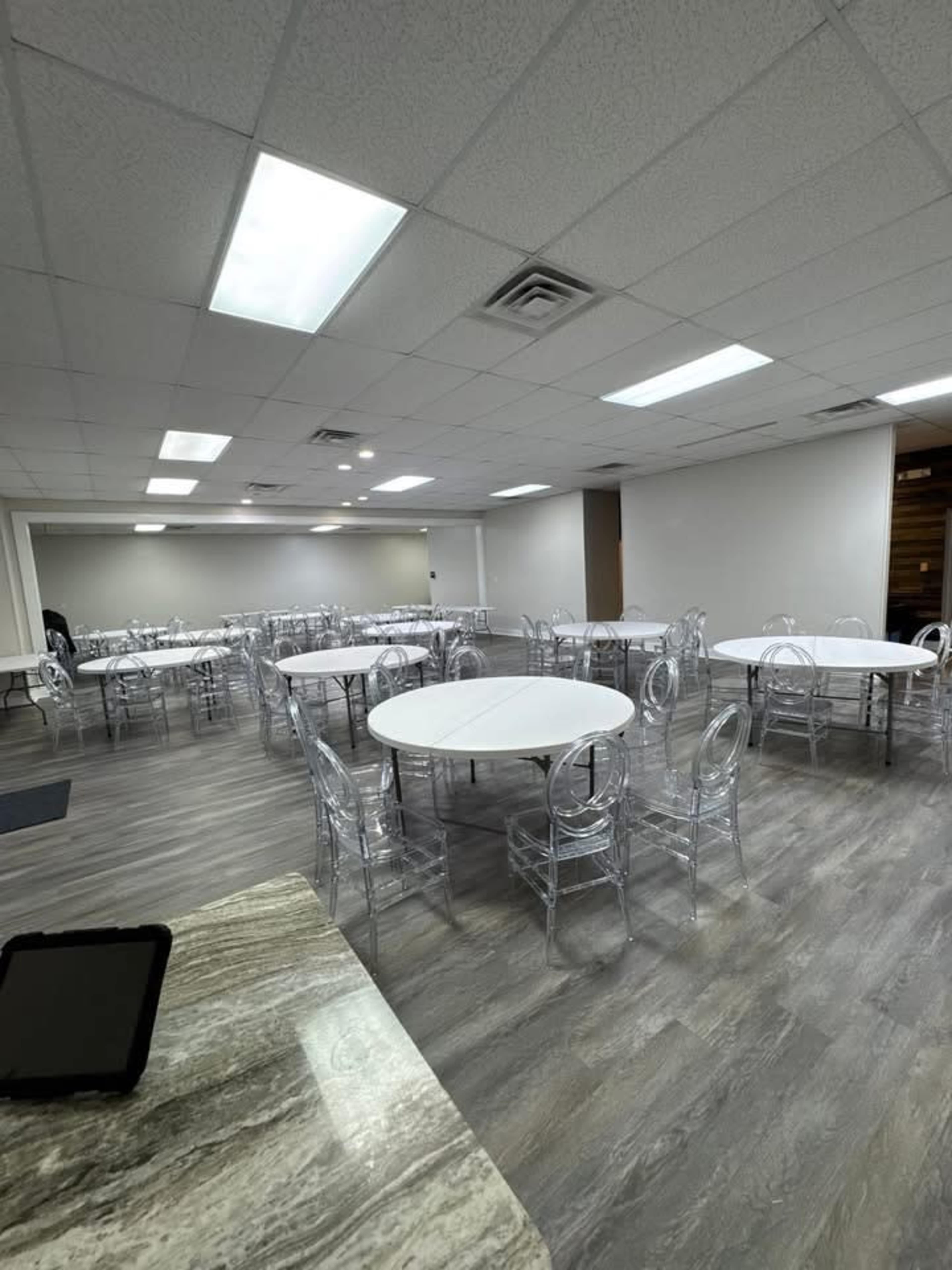 The image shows a spacious room with several round tables set up, each surrounded by transparent acrylic chairs, and equipped with bright overhead lighting.