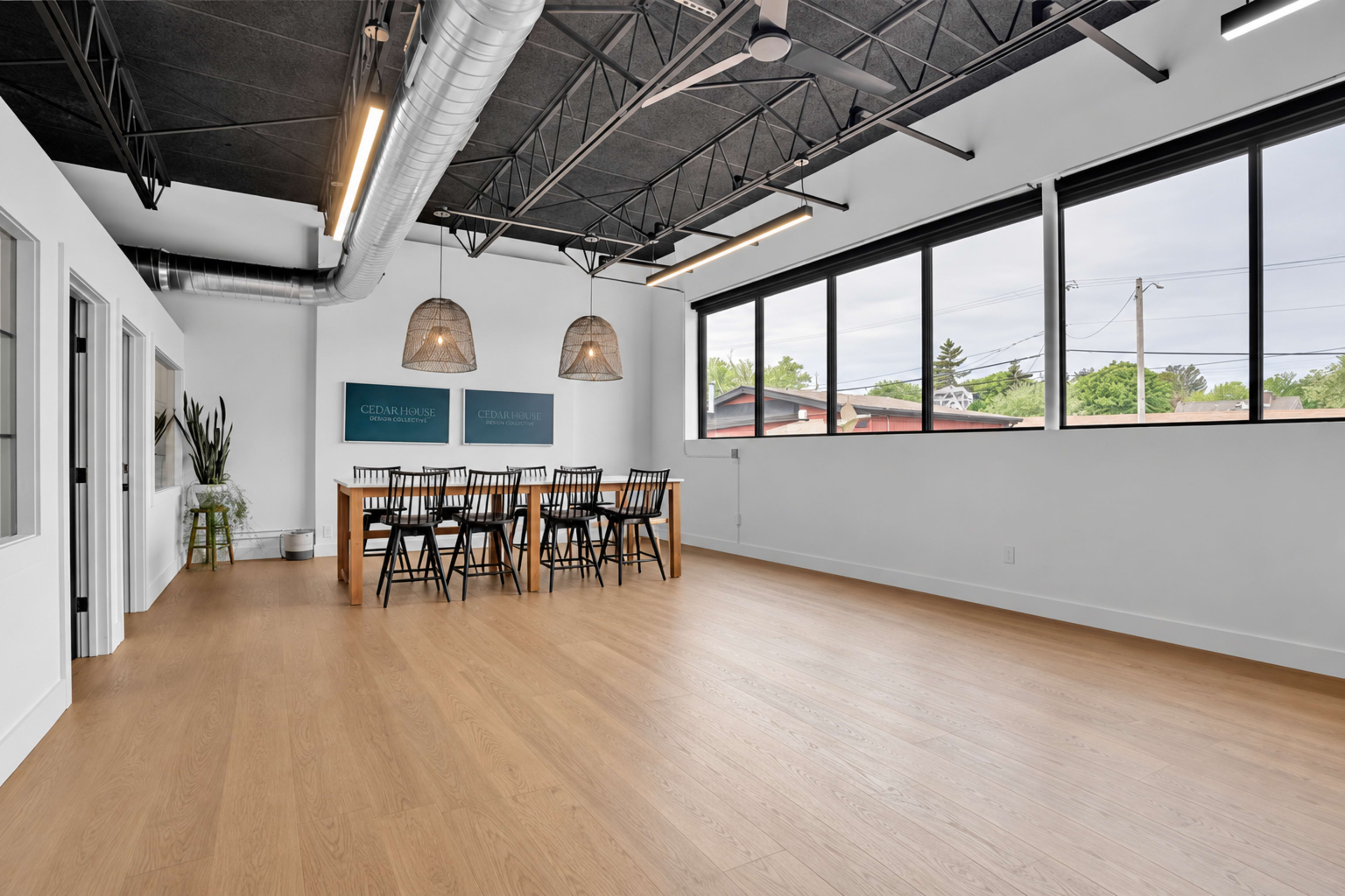 A spacious meeting room features a long wooden table surrounded by chairs, large windows lining one wall, and modern pendant lighting overhead.