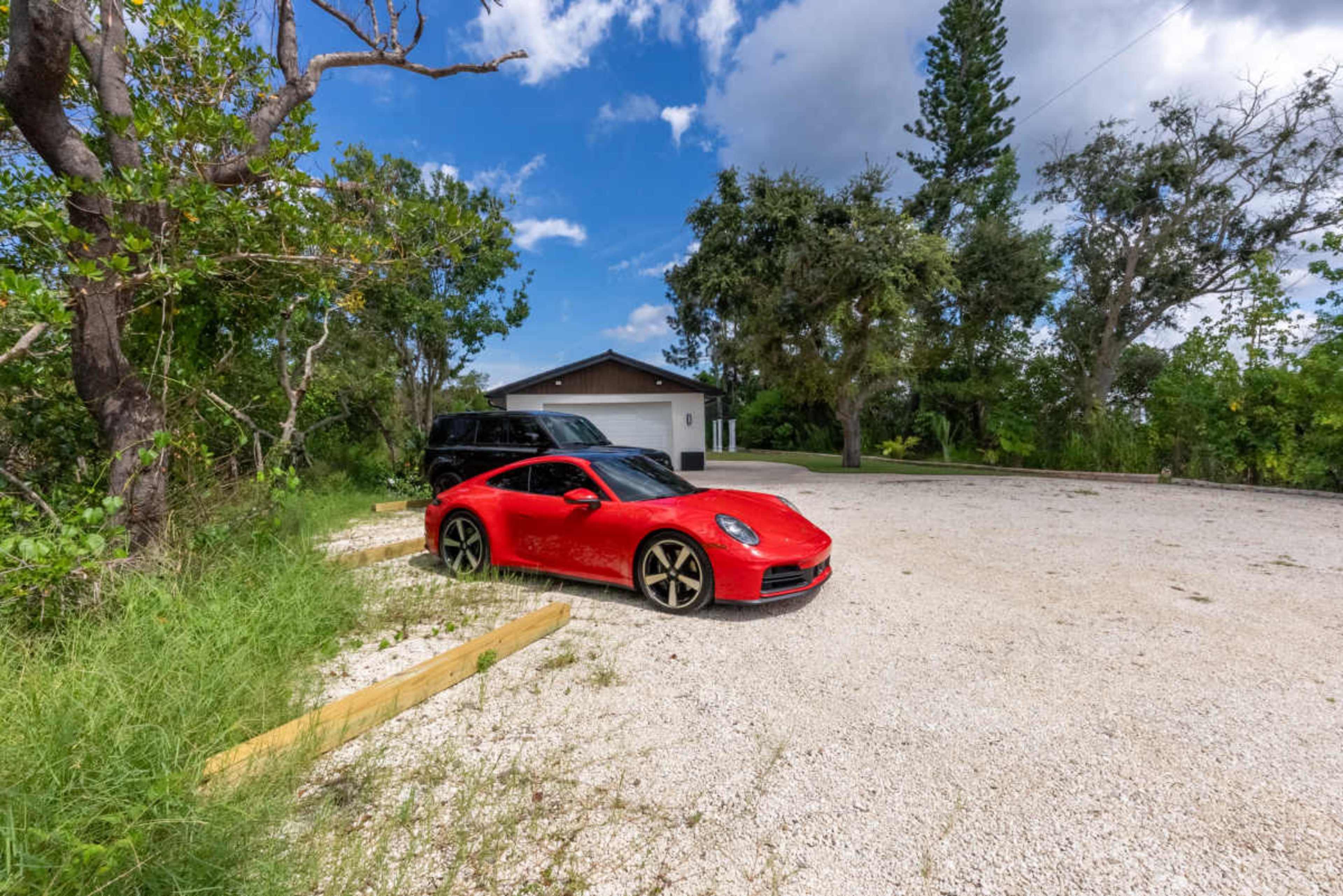 A red sports car is parked on a gravel driveway next to a black SUV, with a house and trees in the background.