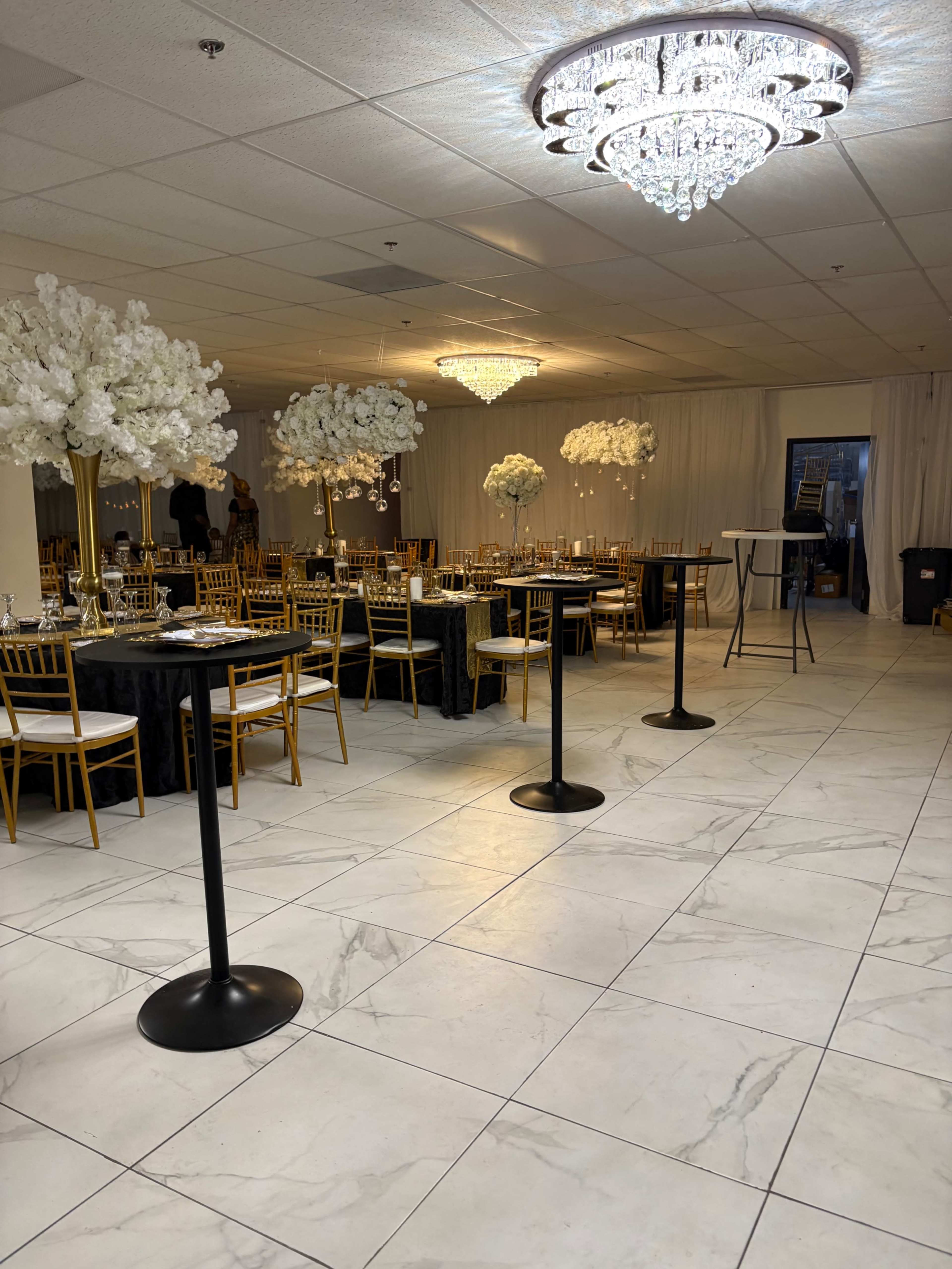 The image shows a banquet hall featuring elegant round tables with black tablecloths, gold chairs, and tall floral centerpieces, illuminated by decorative chandeliers.