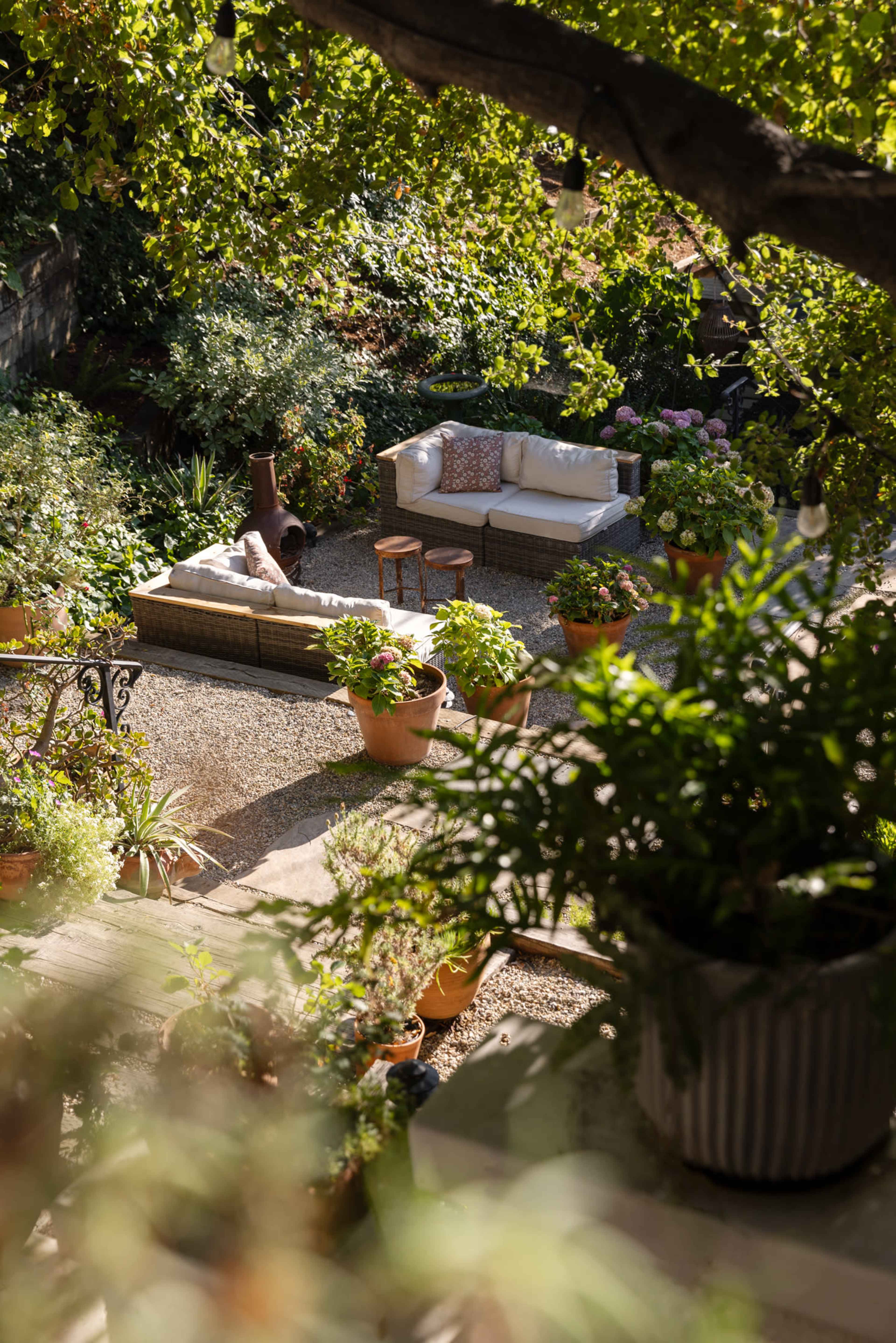 A cozy garden scene features a seating area with cushions, surrounded by potted plants and gravel pathways.