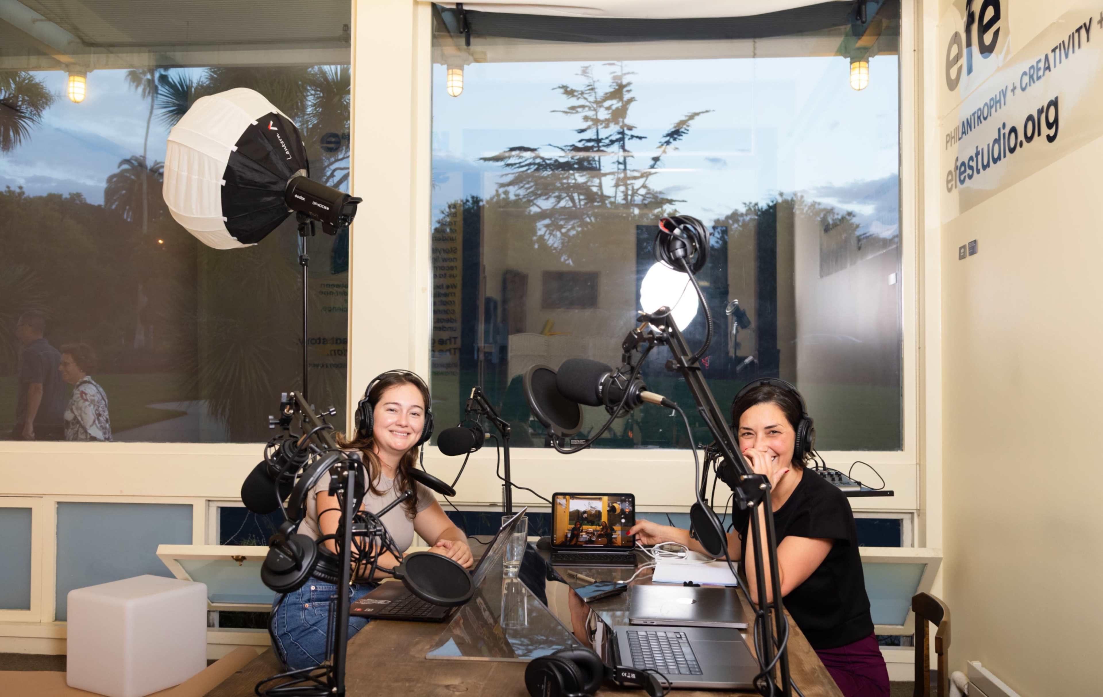 Two women are seated at a table with microphones and recording equipment in a well-lit studio setting with large windows.