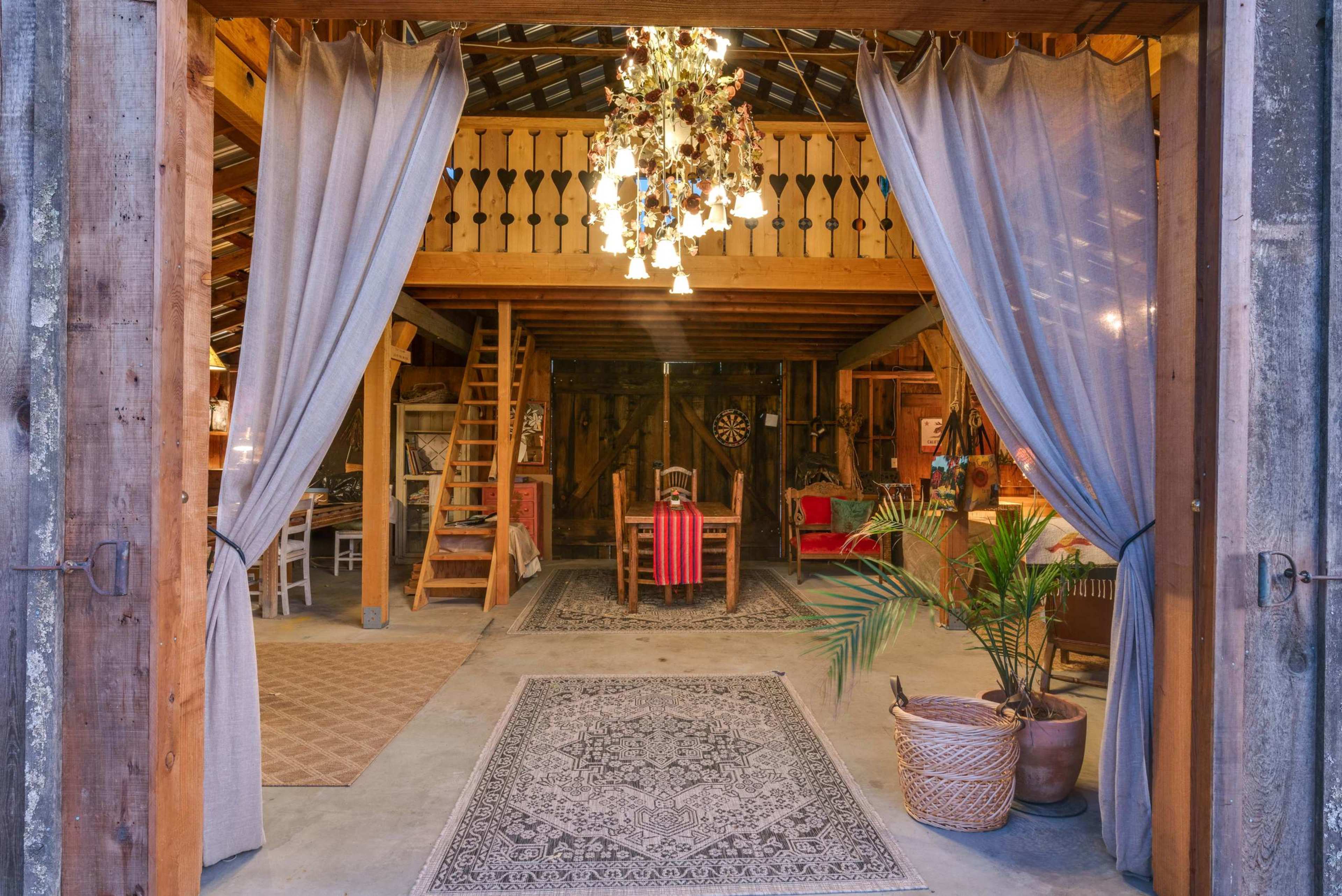 An open barn interior features a dining area with a chandelier, wooden furnishings, and a staircase leading to a loft.