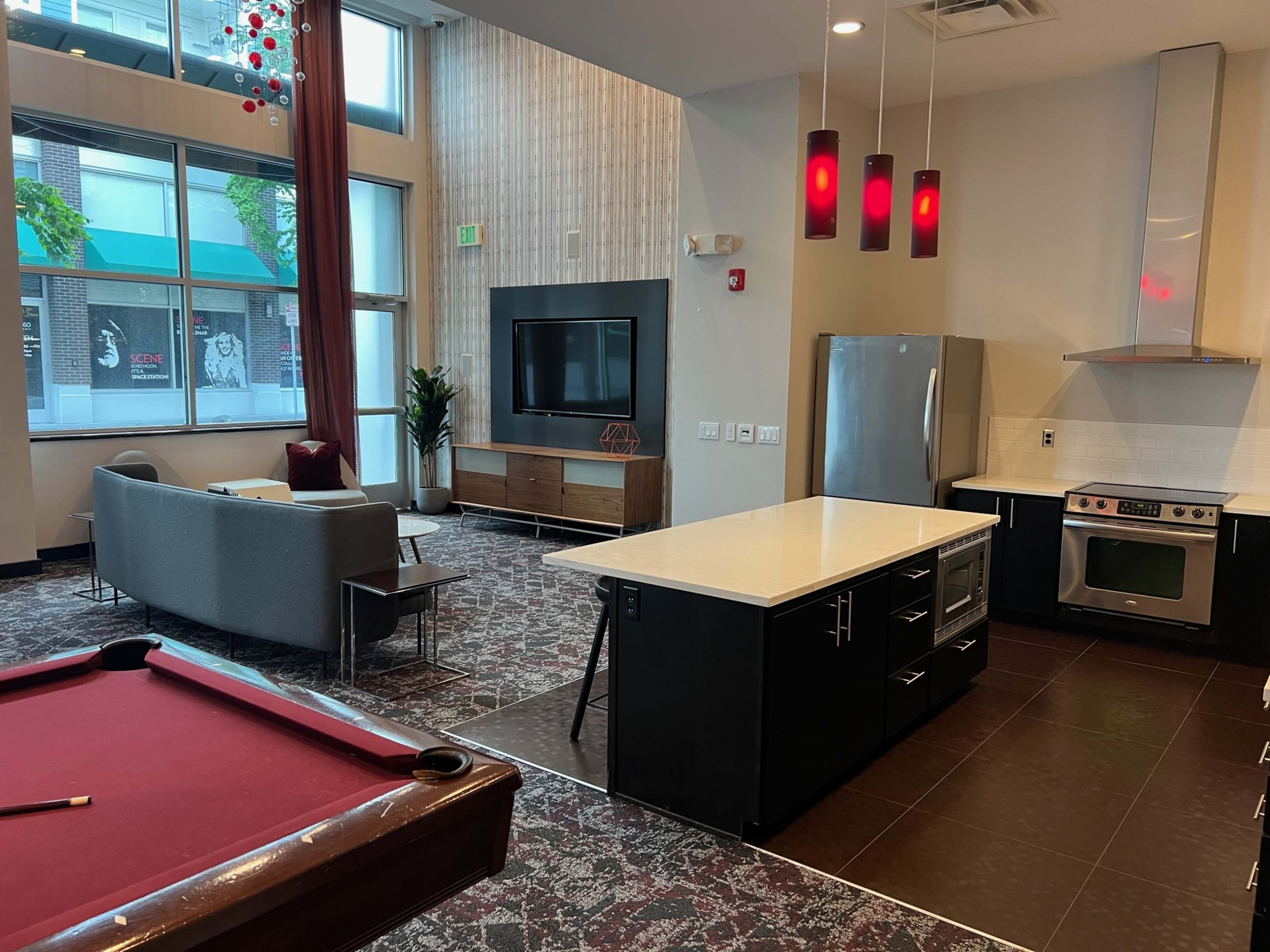 The image shows a modern open-concept lounge area featuring a pool table, a sofa, a kitchen island, and a large TV mounted on the wall.