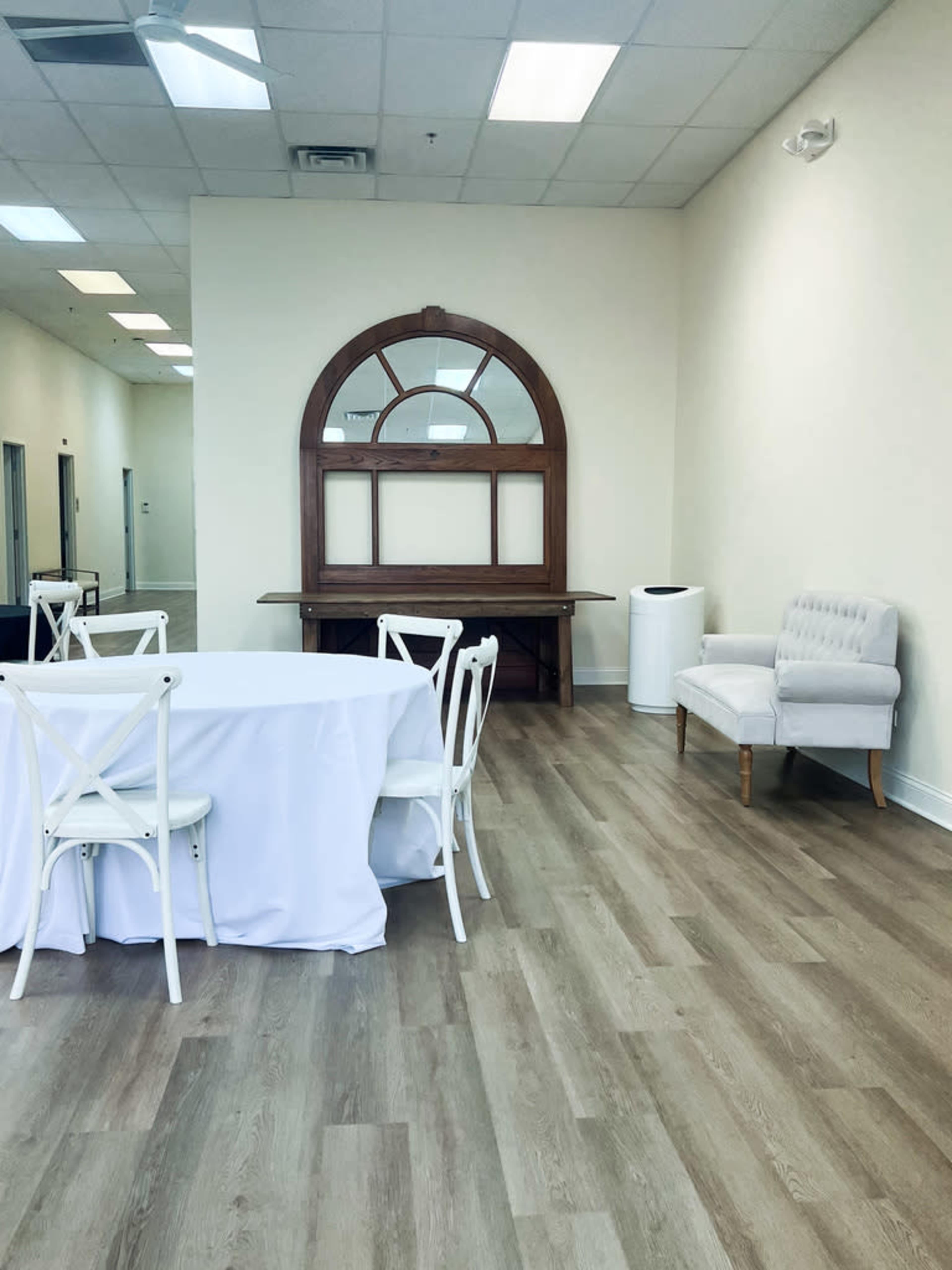 A clean, bright room with a table draped in a white cloth, white chairs, a wooden decorative arch, and a small upholstered chair against the wall.