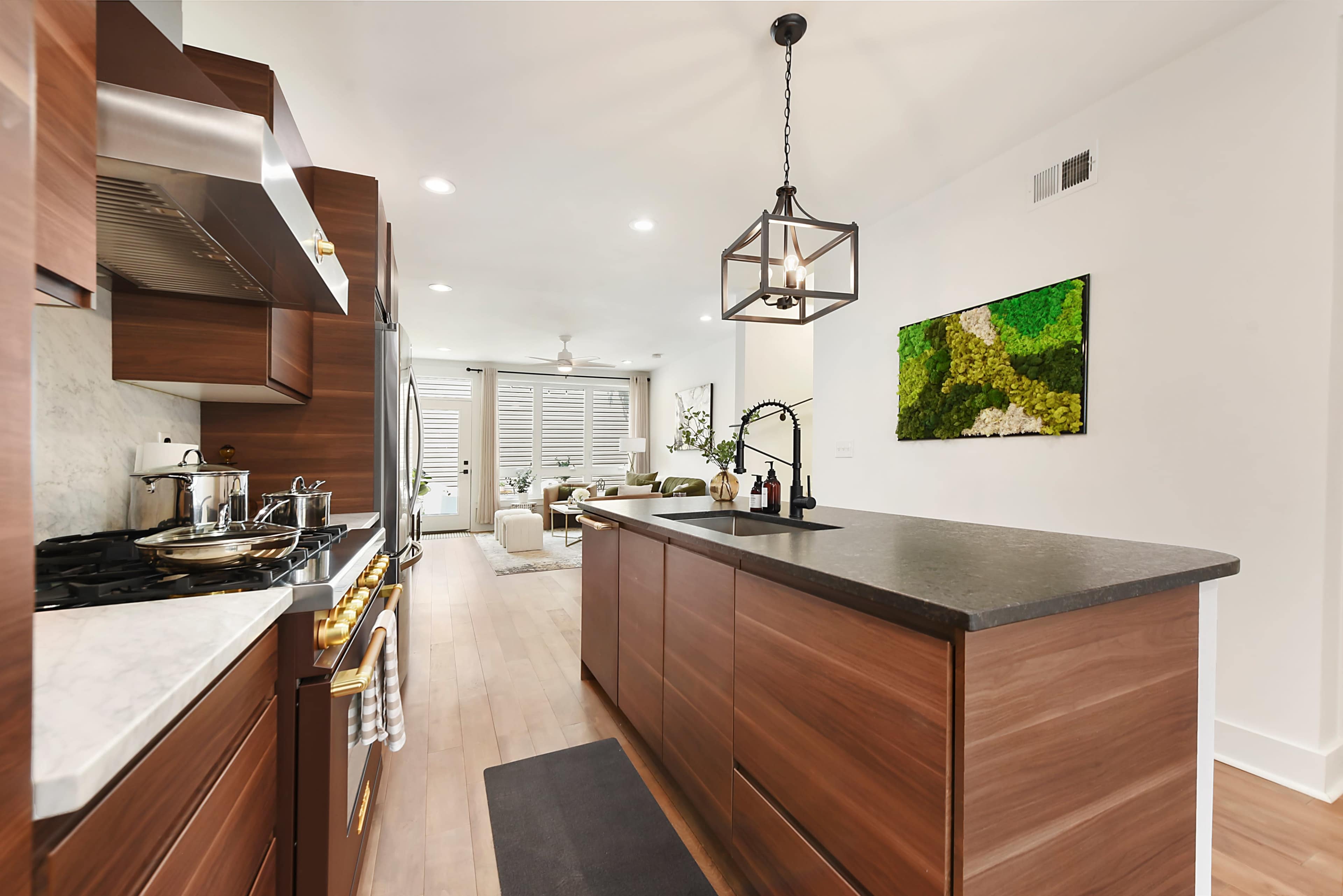 The image shows a modern kitchen with wooden cabinetry, a large island with a dark countertop, and stainless steel appliances, leading into a well-lit living area with greenery.