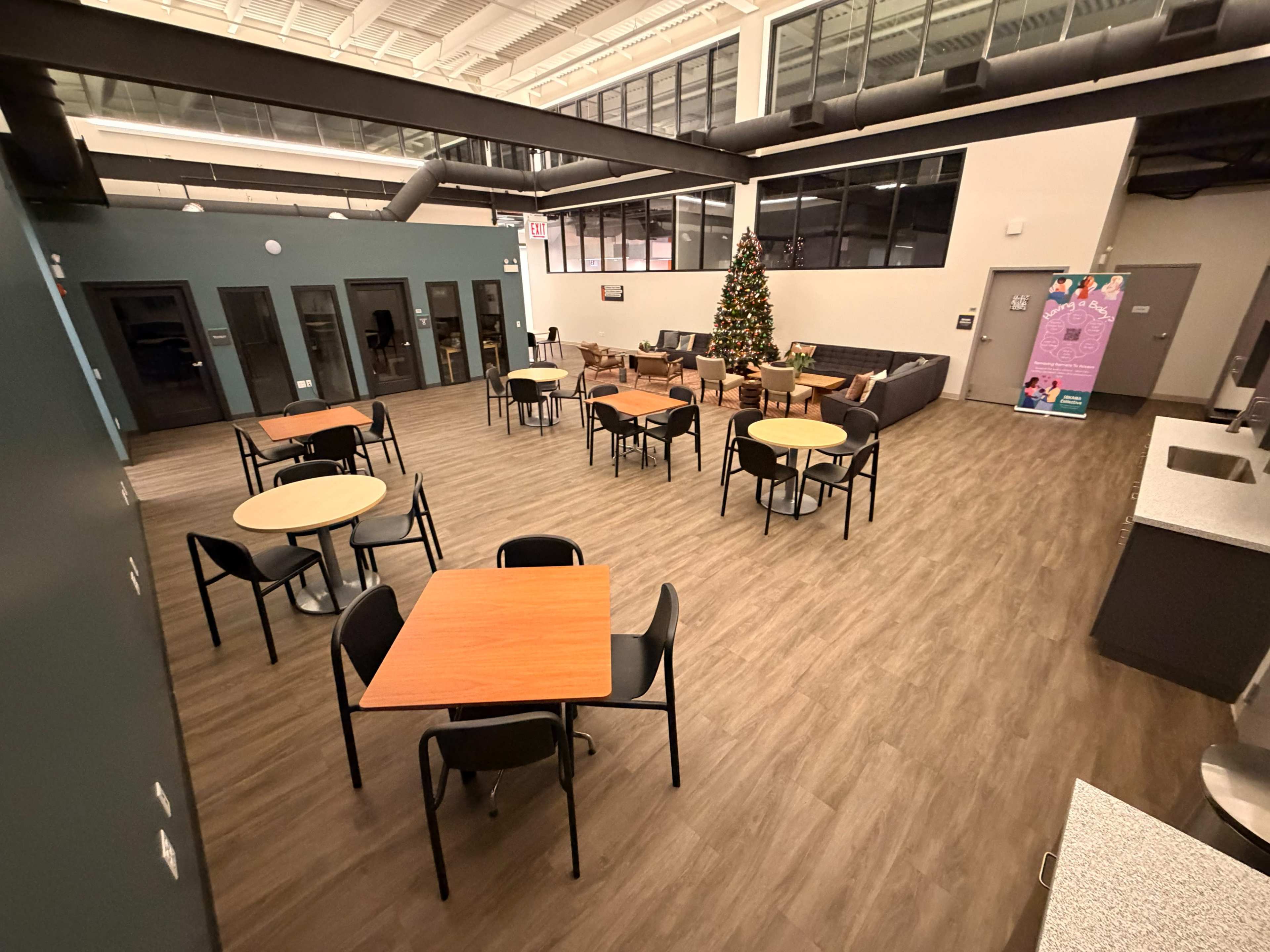 The image shows a spacious indoor area with several tables and chairs arranged, a Christmas tree in the corner, and large windows allowing natural light.
