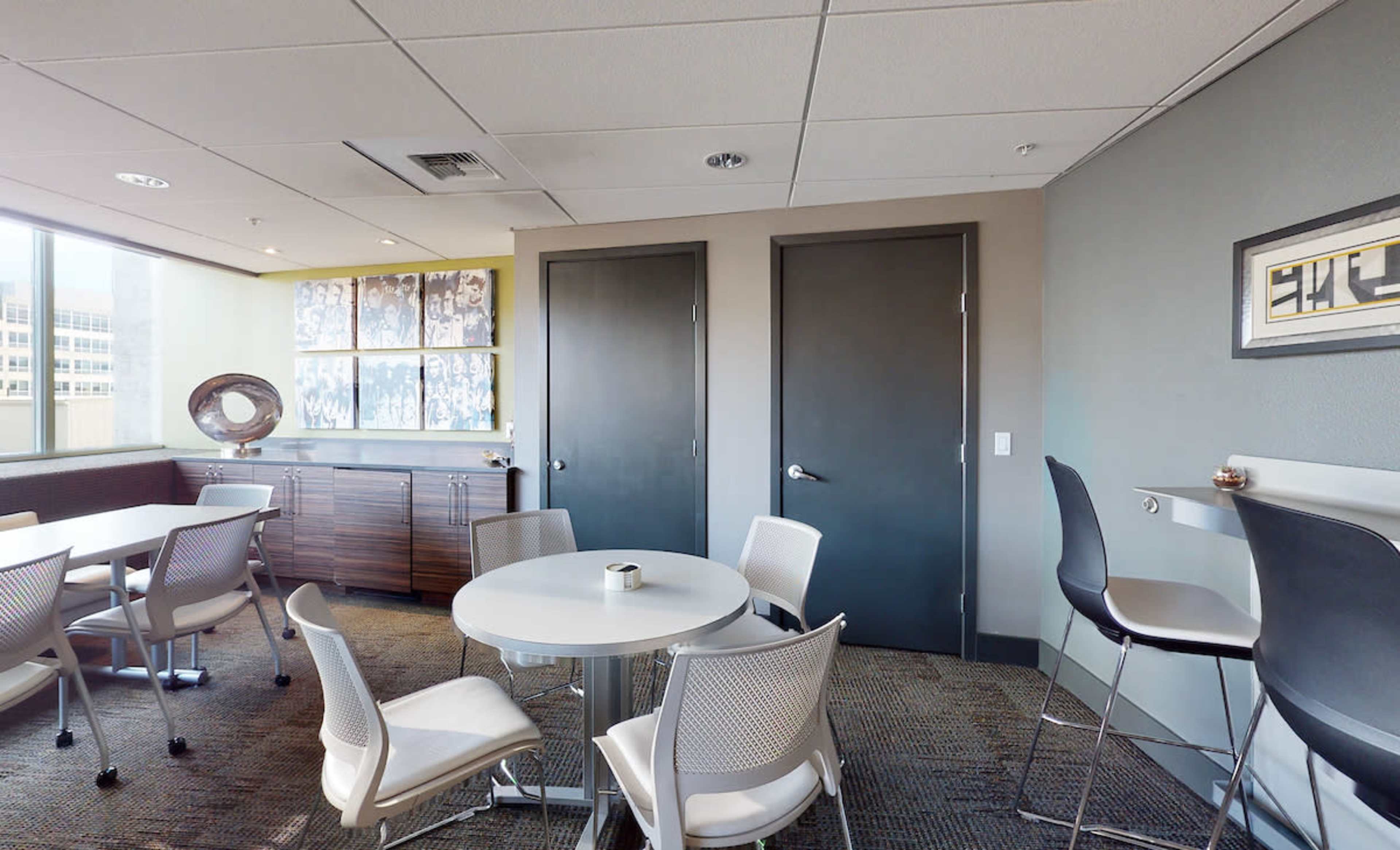 A modern office lounge features a round table surrounded by white chairs, with two closed doors and a side cabinet in the background.