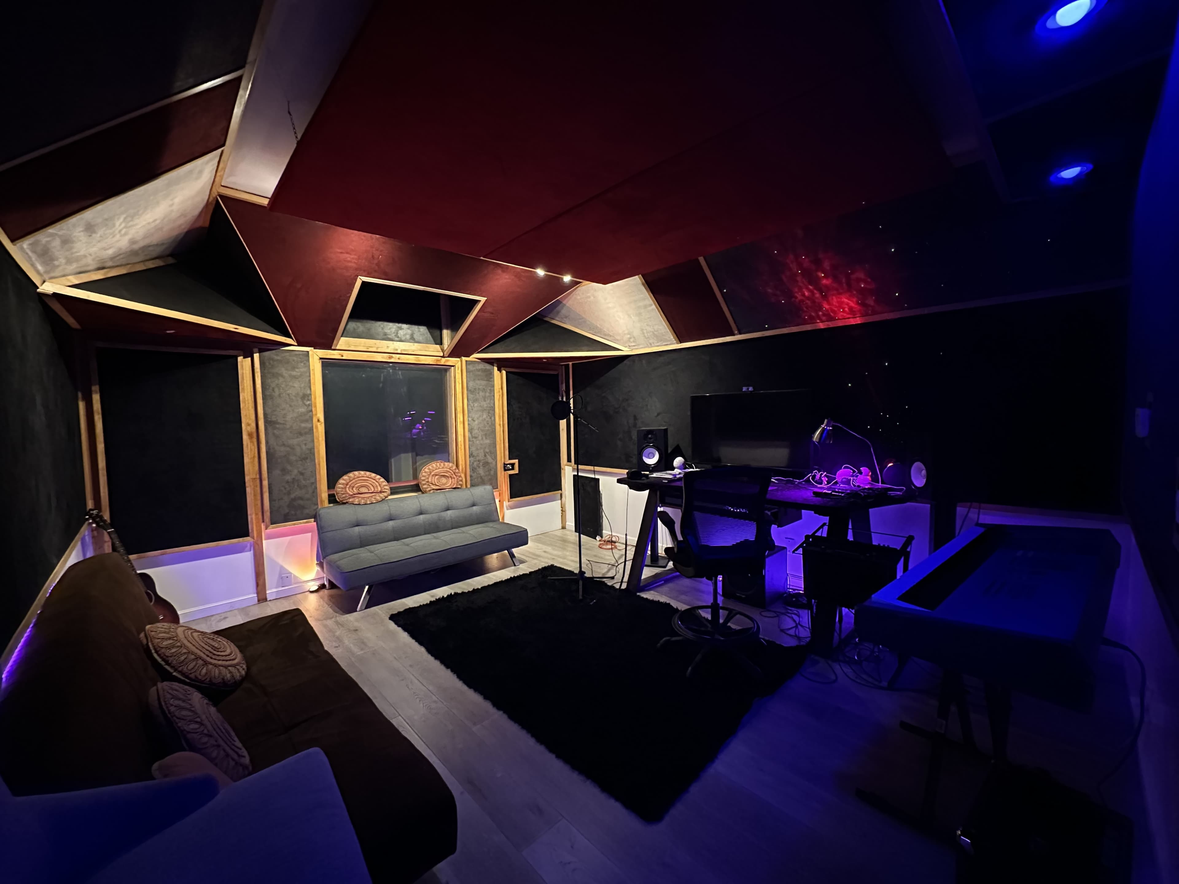 A music studio with a sectional couch, a desk with a computer and audio equipment, and colorful lighting on the ceiling.
