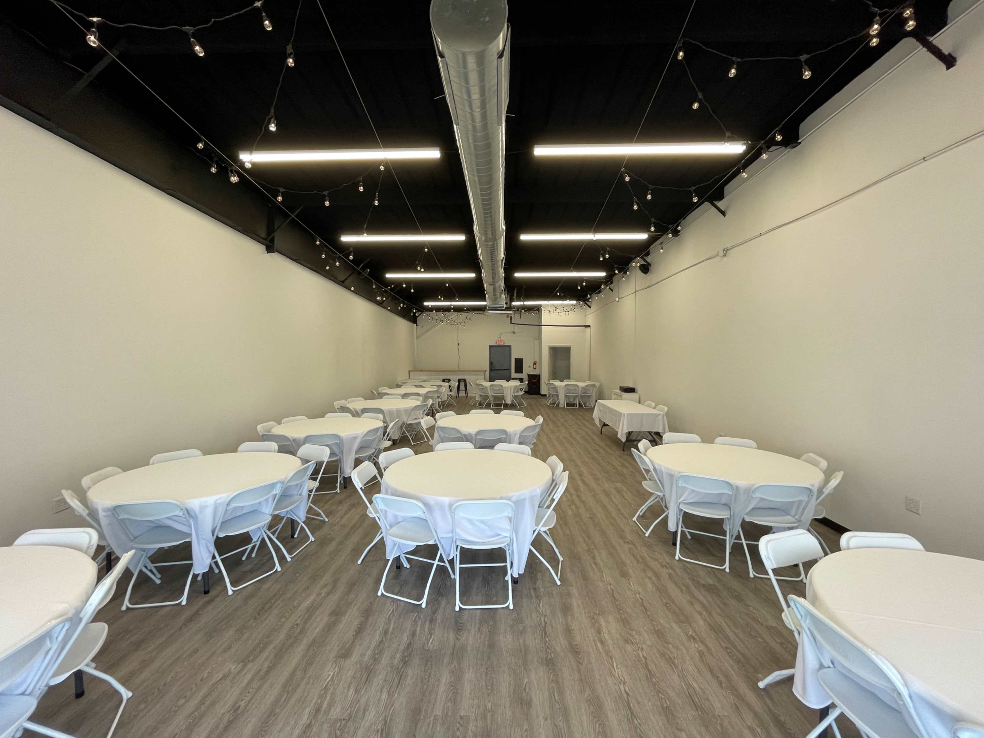 A large, well-lit event space with several round tables covered in white tablecloths and arranged neatly across the floor.