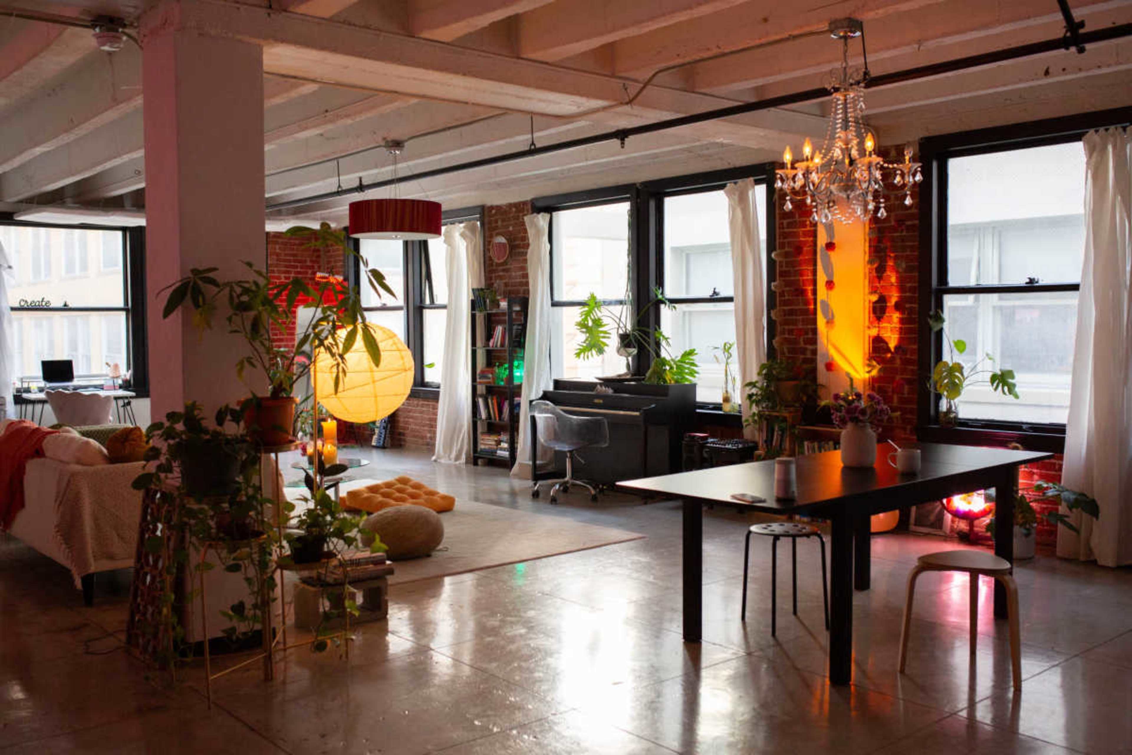 The image shows a spacious open-concept living area with large windows, a dining table, plants, and various cozy furnishings.