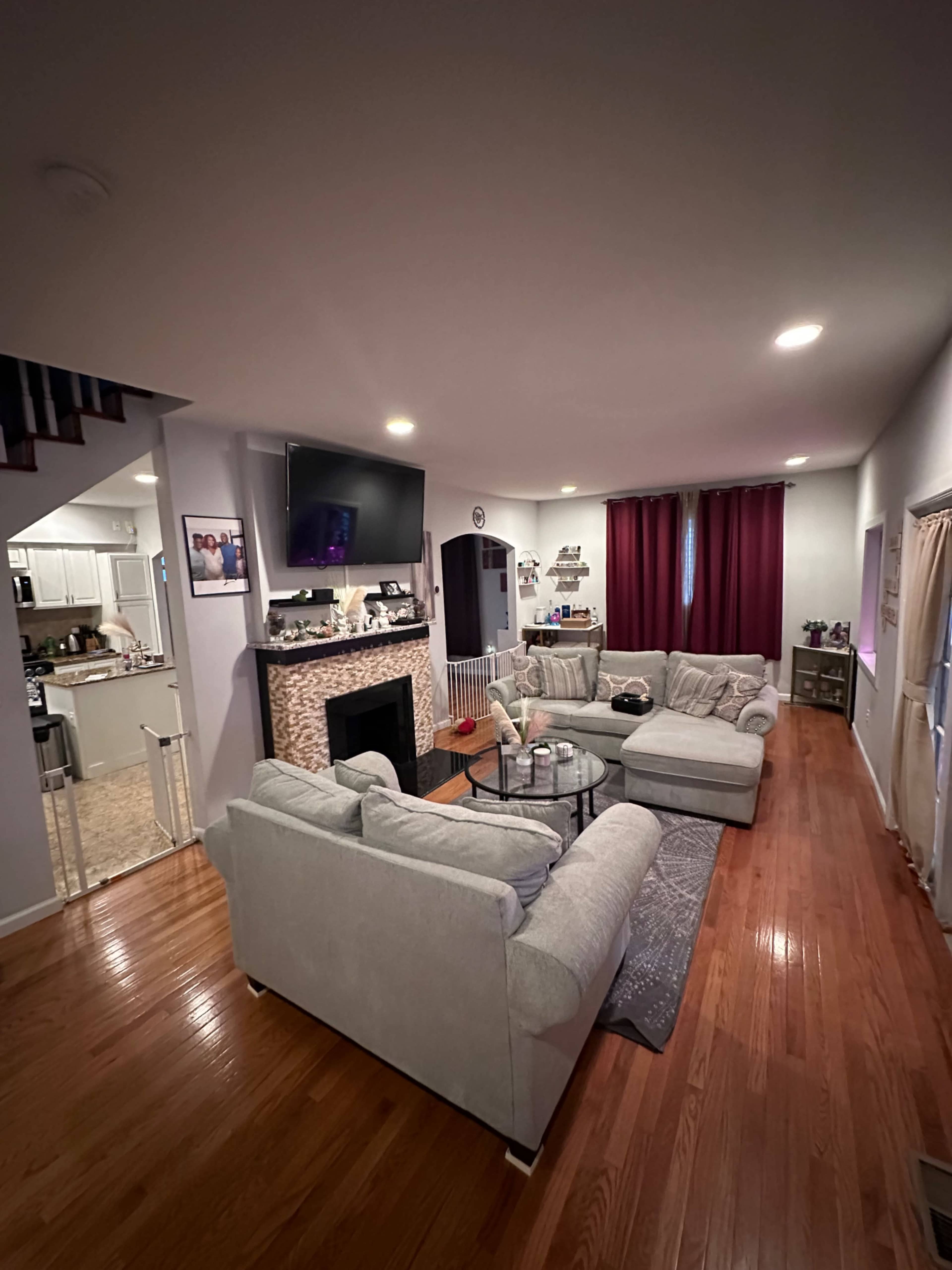 The image shows a modern living room with a beige sectional sofa, a coffee table, a TV mounted above a stone fireplace, and red curtains on the windows.