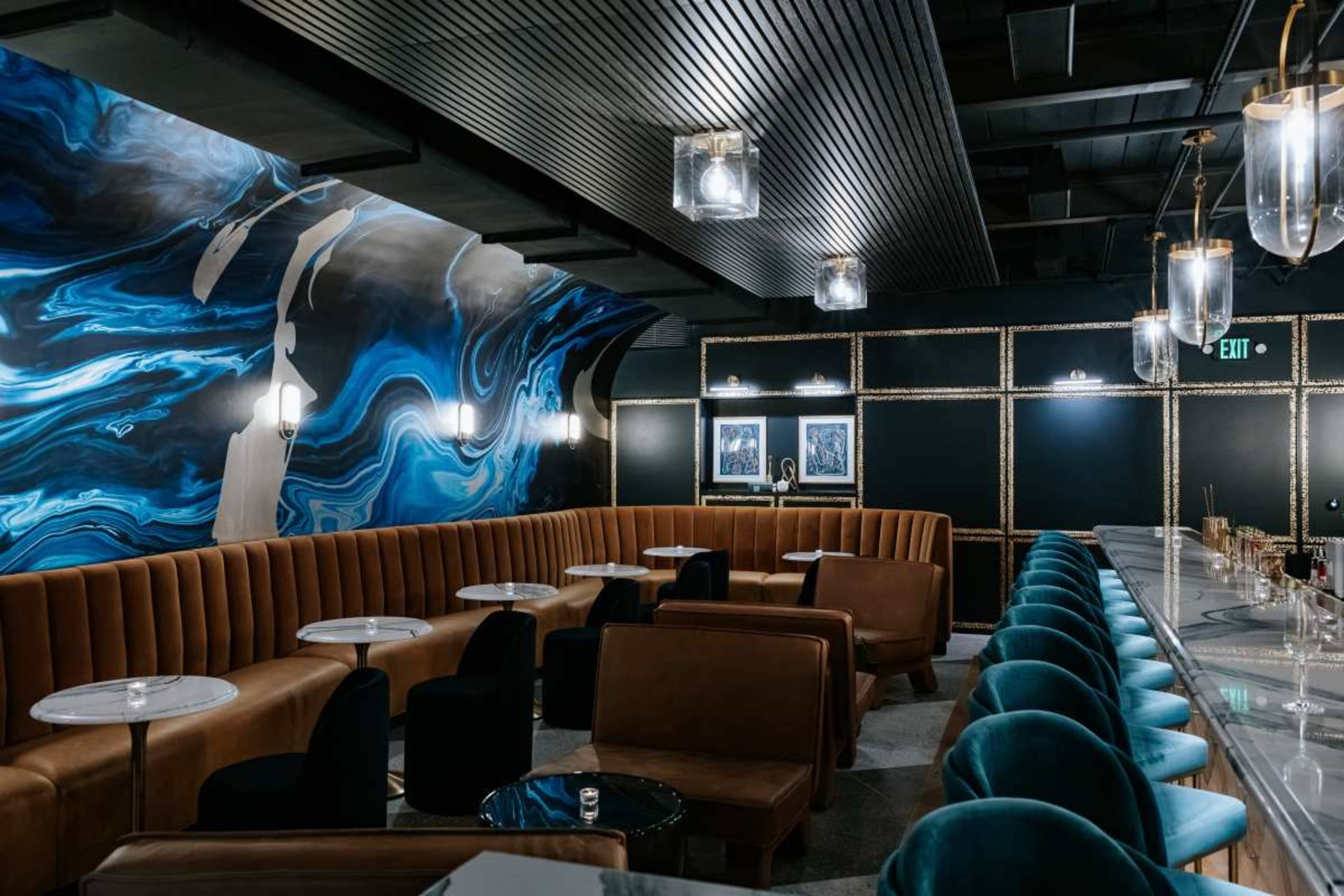 The image shows a modern lounge with brown leather seating, blue accents on the walls, and a long bar with glowing pendant lights.