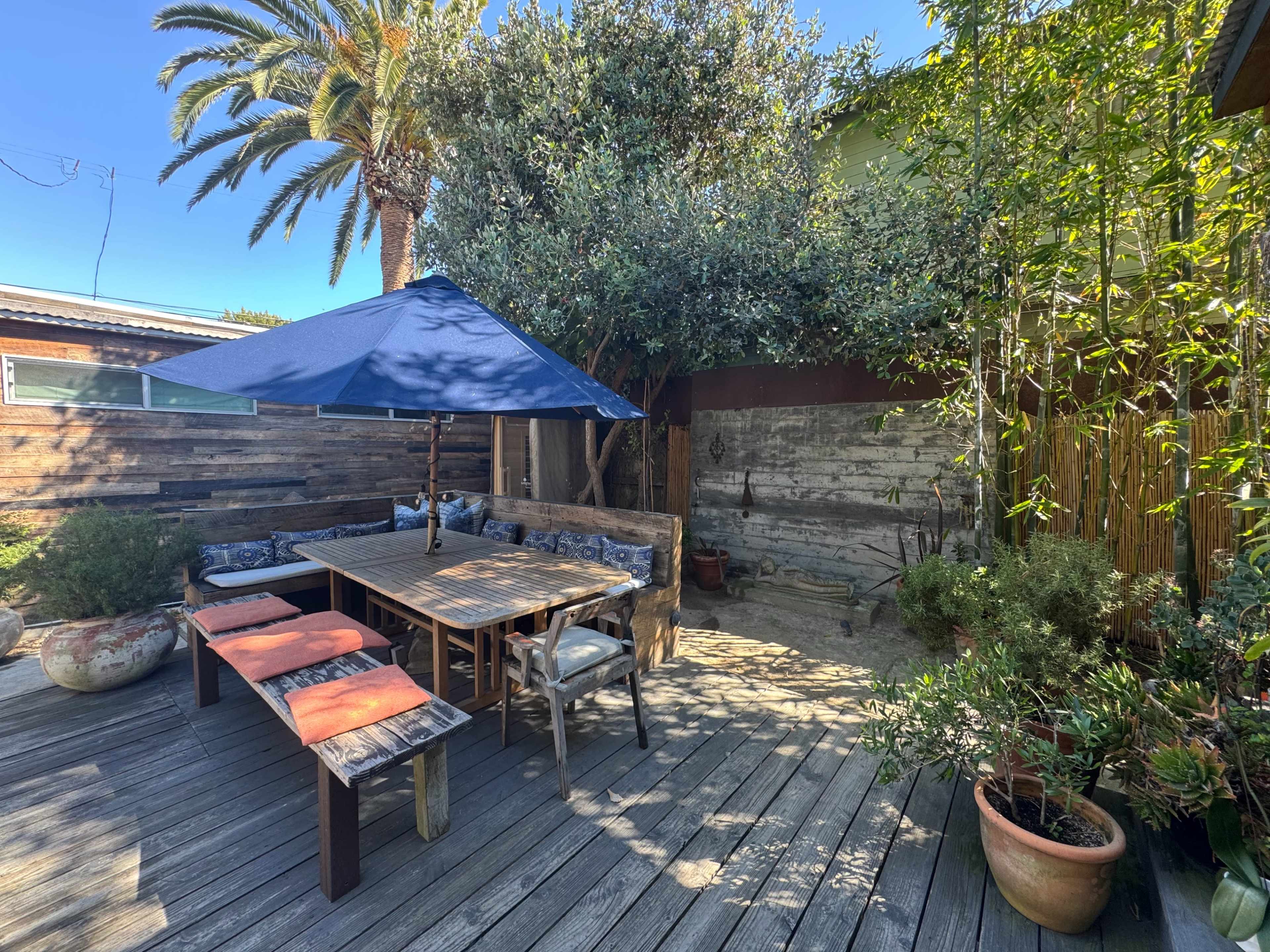A wooden patio features a table with seating under a blue umbrella, surrounded by greenery and palm trees.