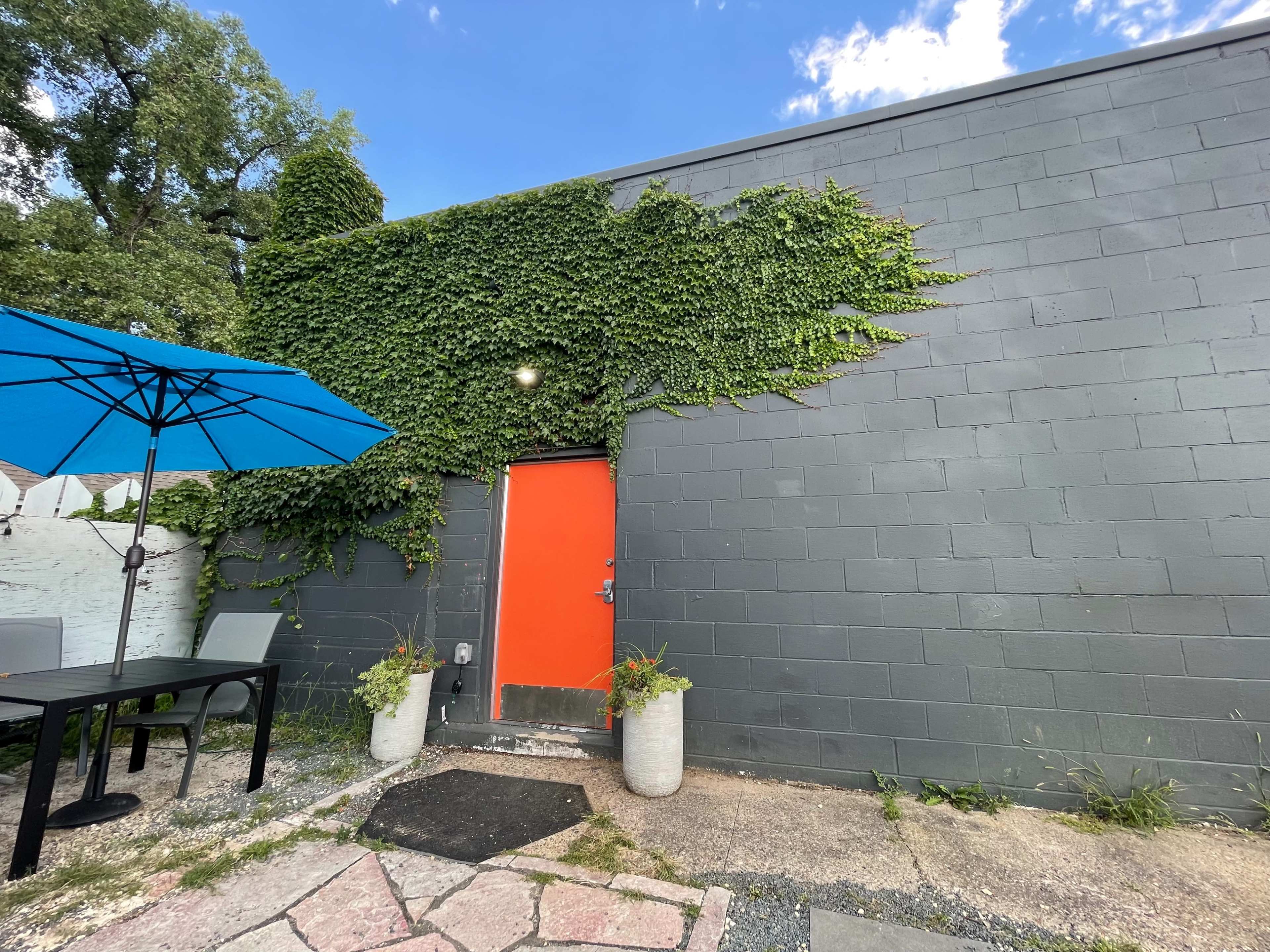 A gray wall with green ivy on it features a bright orange door, surrounded by a stone pathway and outdoor furniture under a blue umbrella.