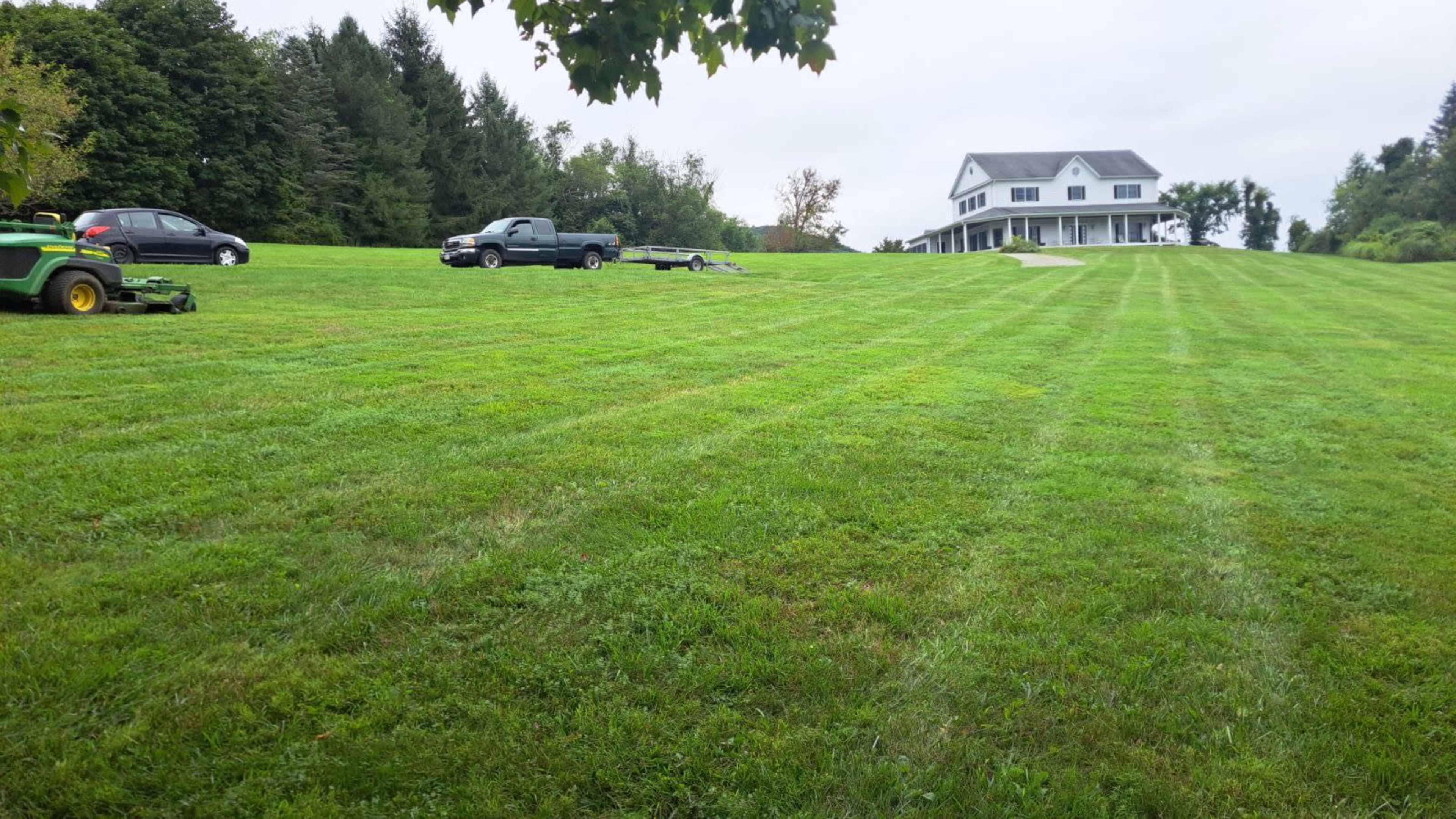 A well-maintained lawn with freshly mowed stripes stretches down to a large two-story house in the background, accompanied by several parked vehicles.