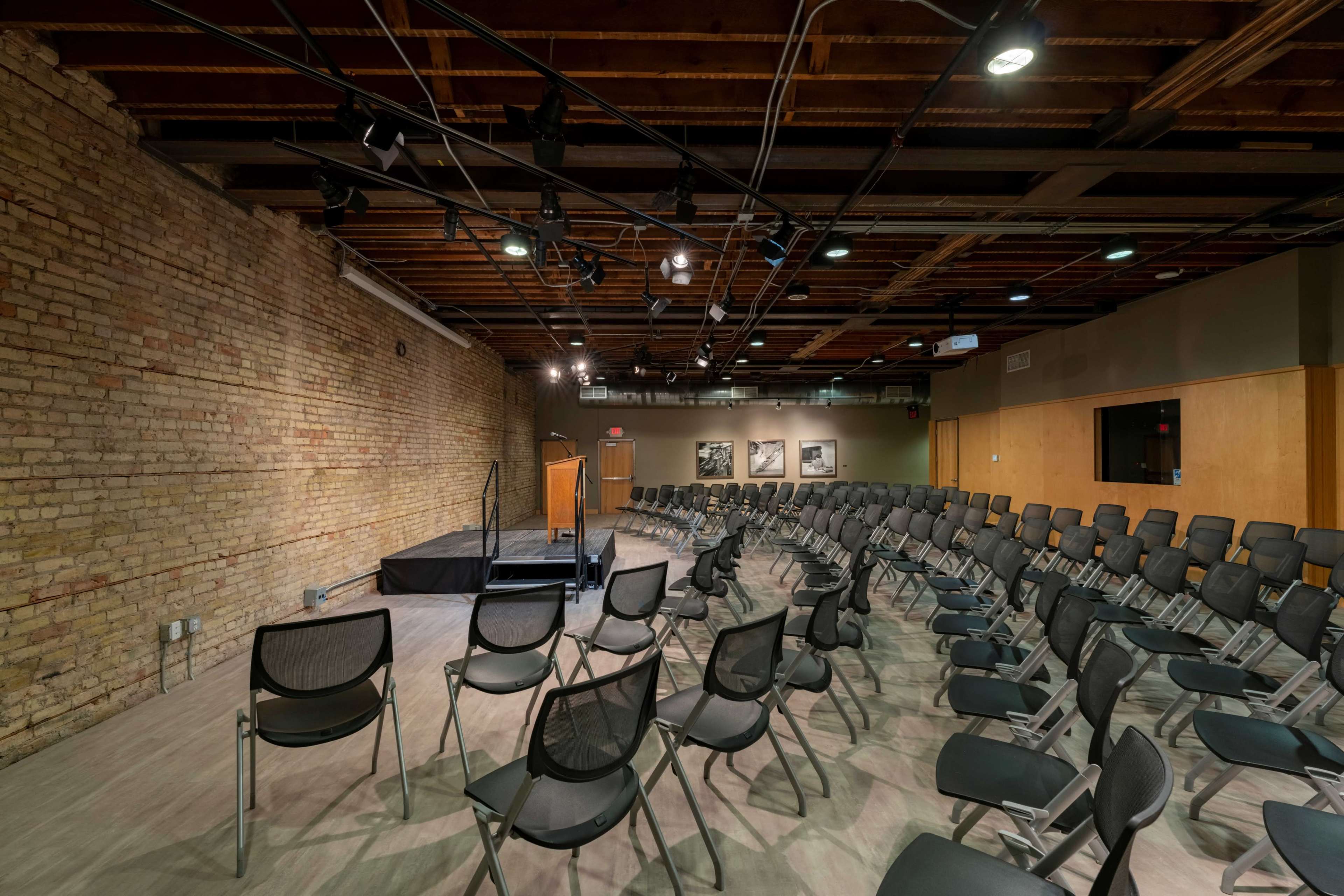 An empty auditorium with rows of black chairs facing a small stage against a brick wall.