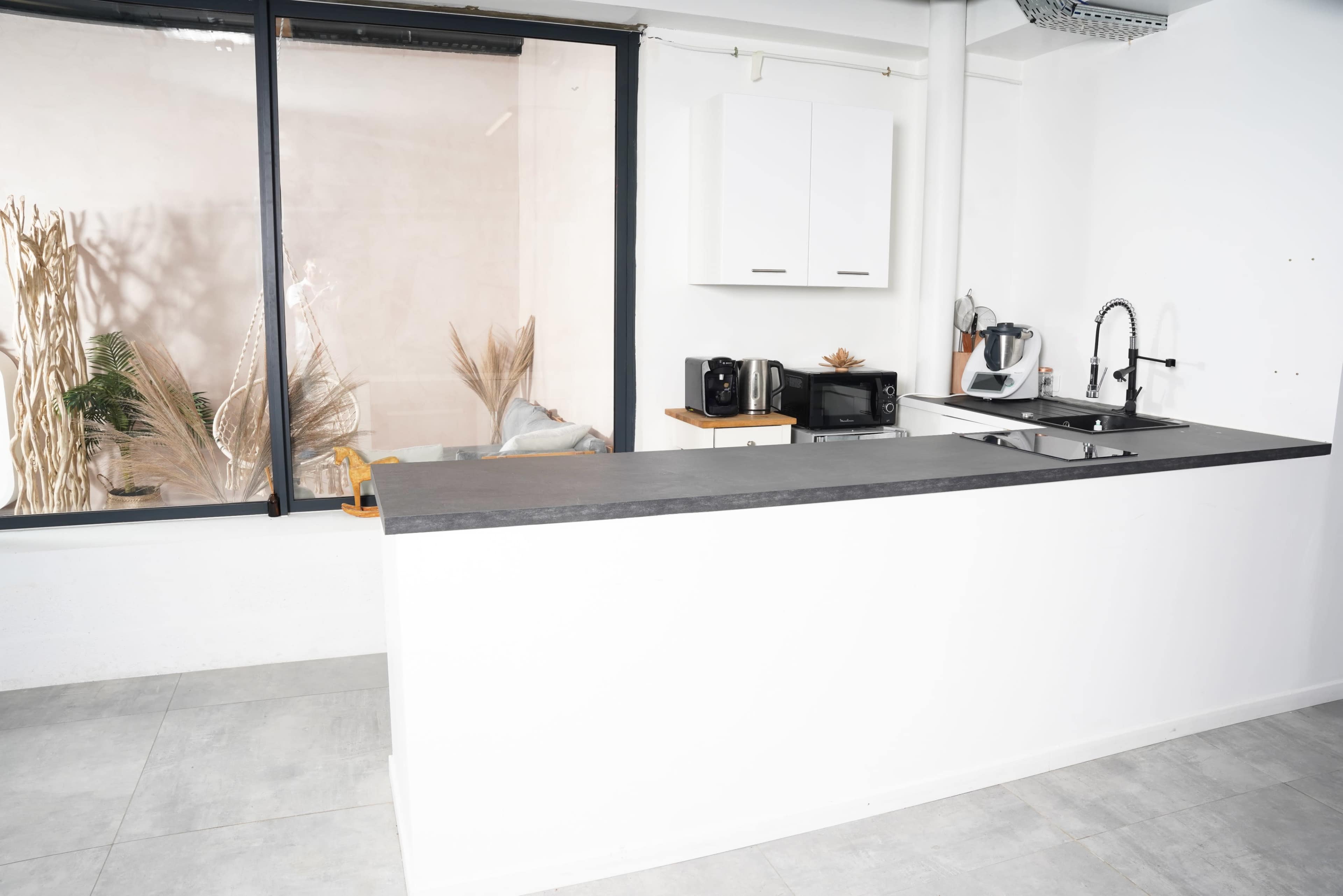 A modern kitchen with a white bar counter, a sink, and small appliances set against a large window displaying plants in the background.