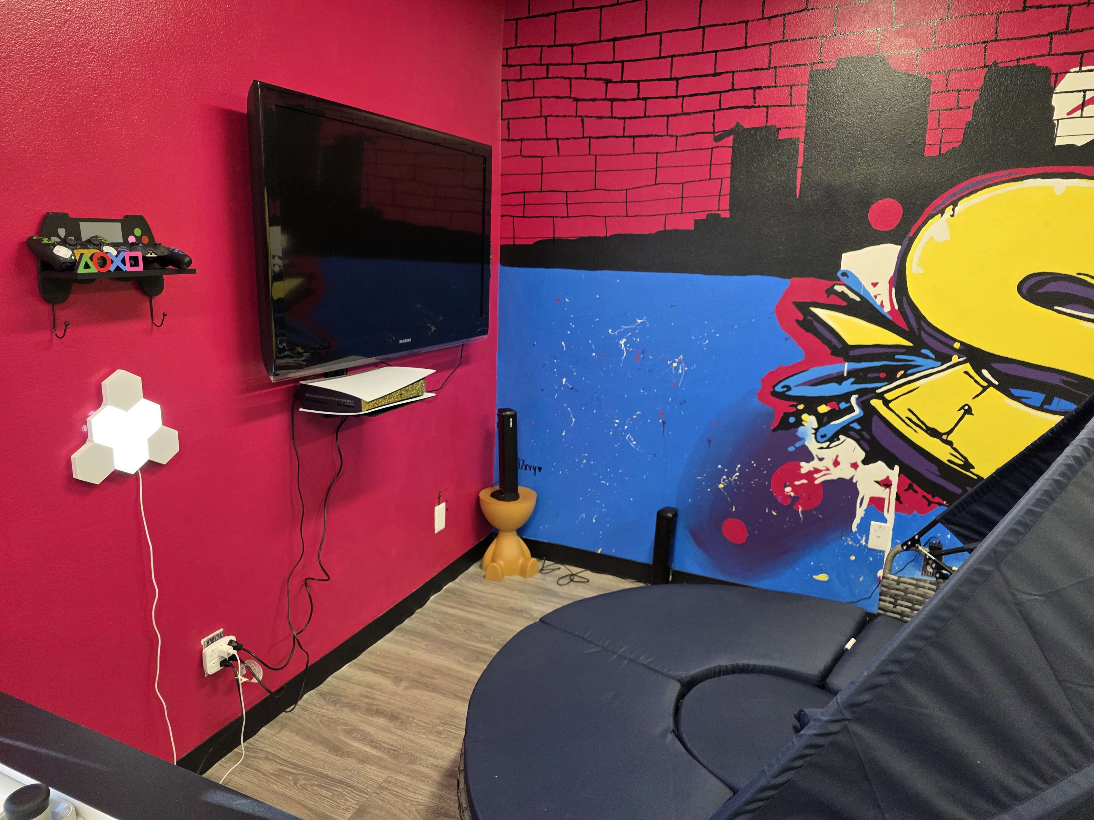 The image shows a gaming setup with a television mounted on a red wall featuring graffiti art, a gaming console below it, a controller on a shelf, and a circular blue gaming chair in the foreground.