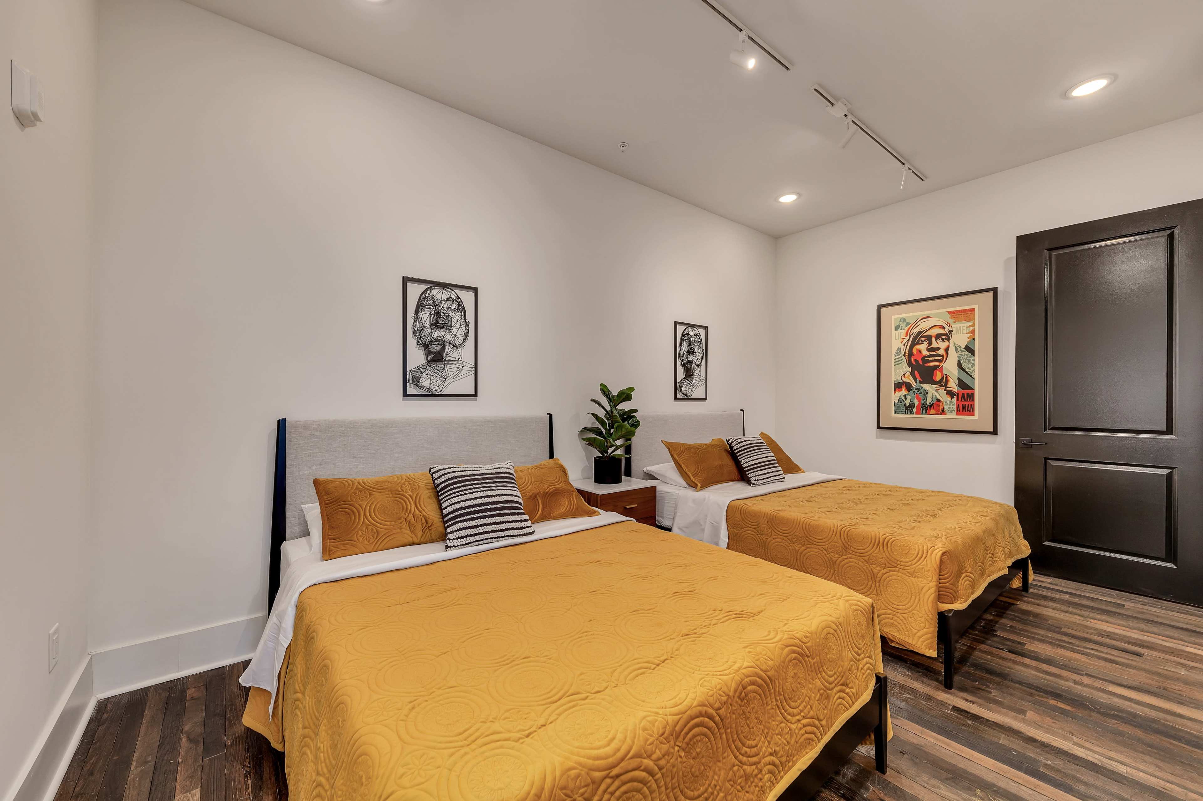 The image shows a modern bedroom with two beds covered in yellow quilts, a nightstand between them, and framed artworks on the walls.