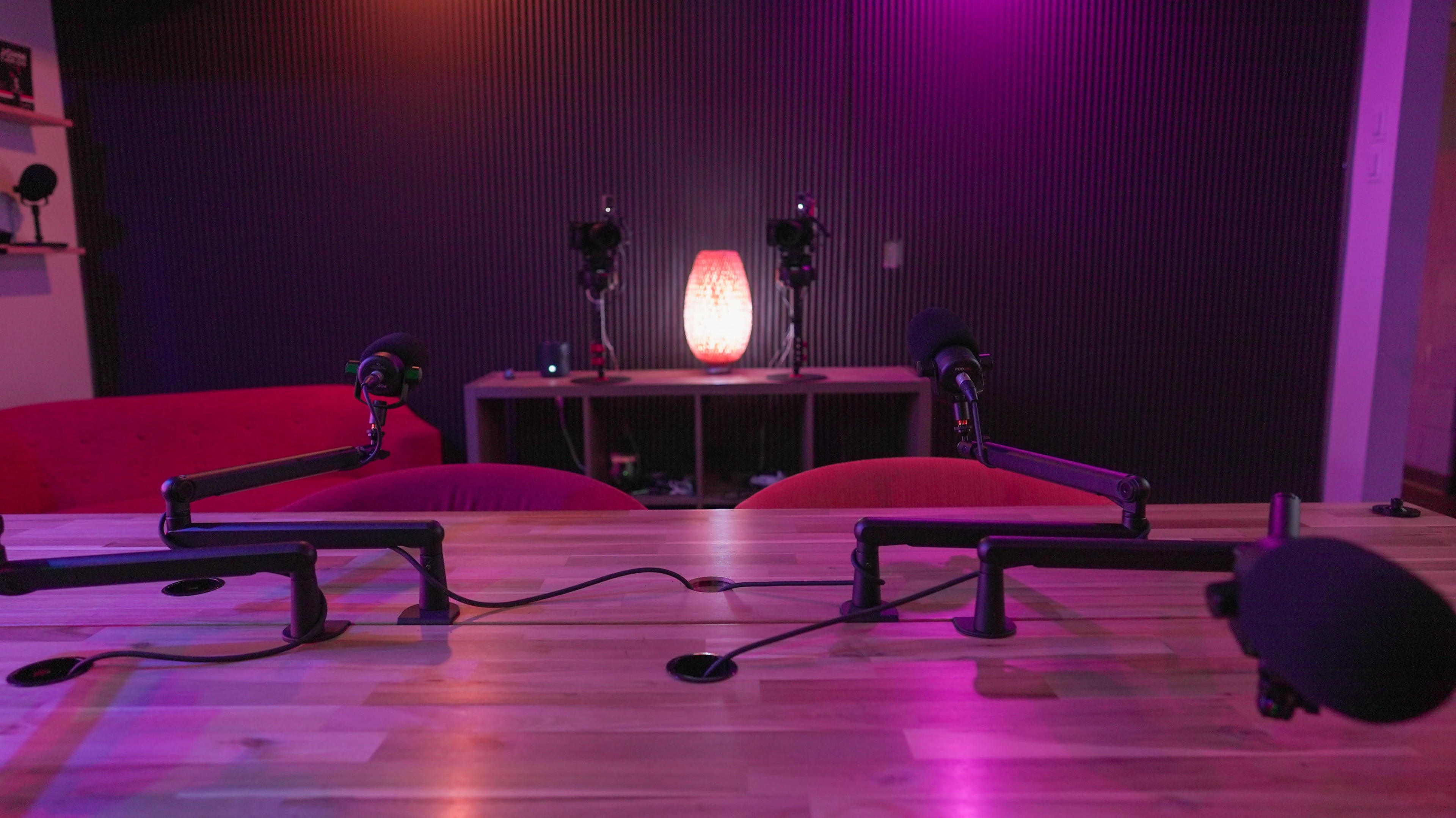 A podcasting setup with two microphones positioned on a wooden table, facing a soft-lit lamp and shelves in a dimly lit room.