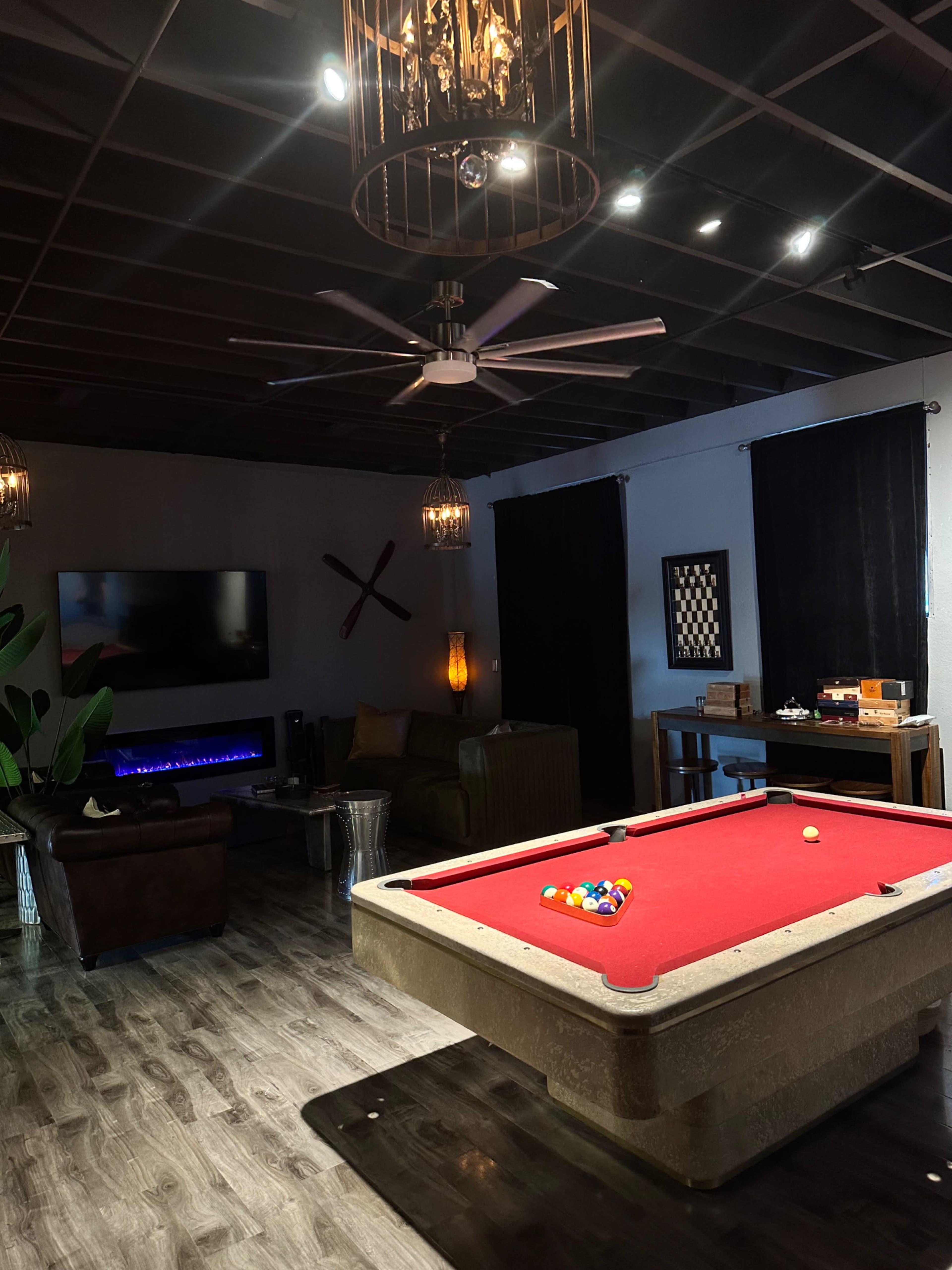 The room features a pool table at the center, surrounded by a seating area, a large television, and decorative elements like a chessboard and plants.