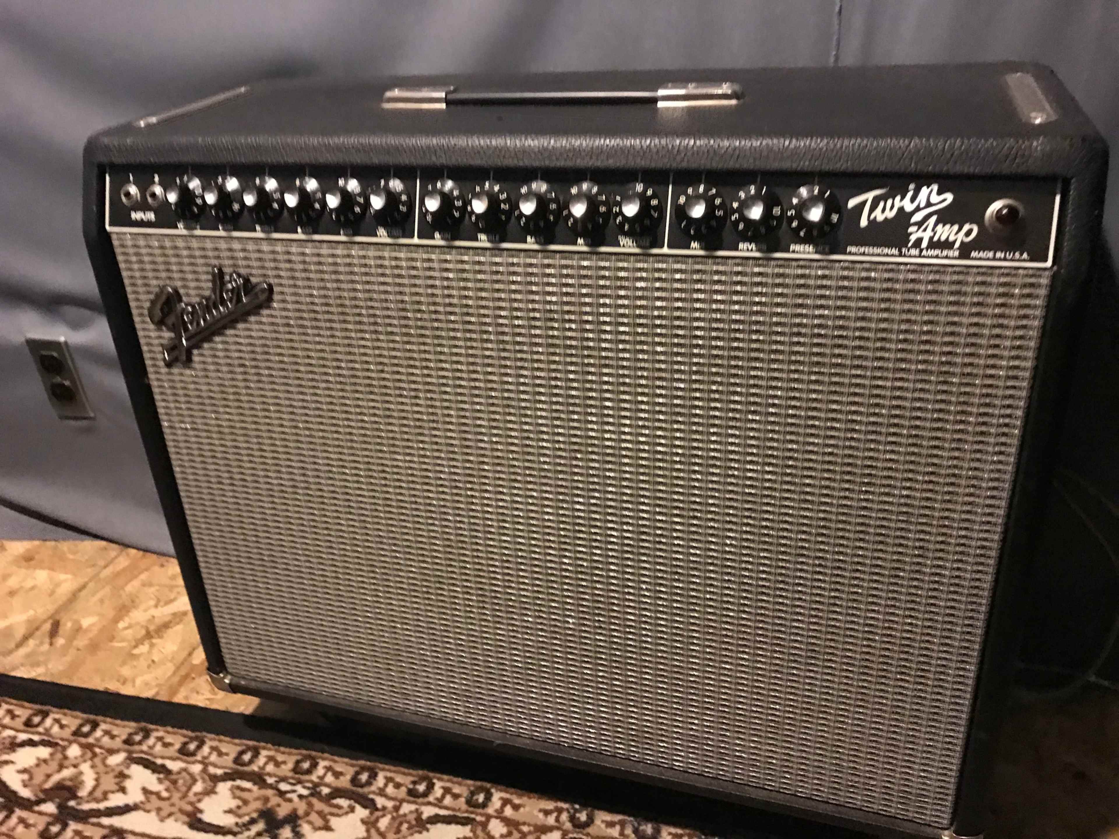 The image shows a black Fender Twin Amp guitar amplifier with multiple knobs on its front panel, placed on a patterned rug against a gray backdrop.