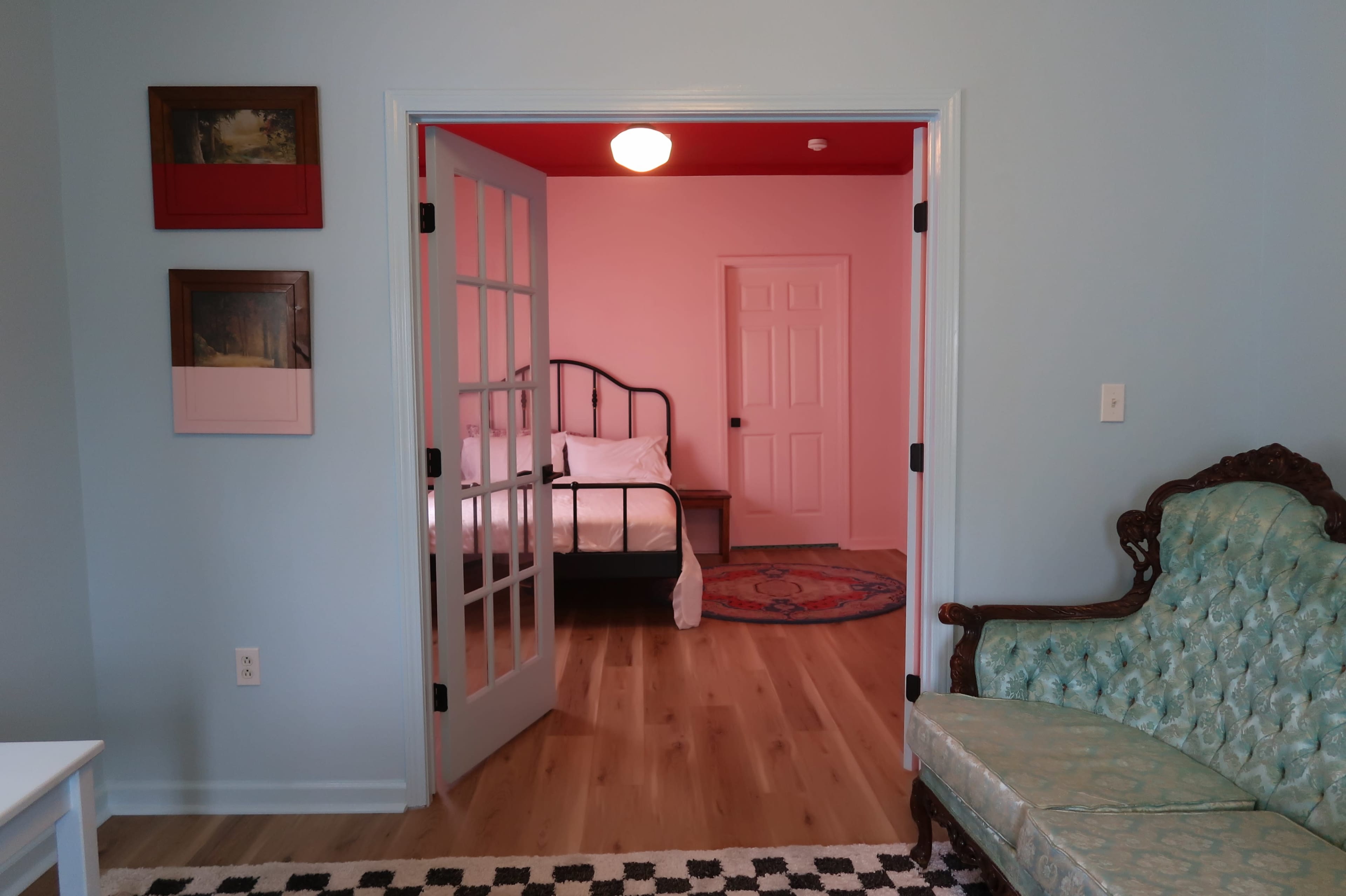 A light blue room featuring a green upholstered sofa, framed artwork on the wall, and an open doorway leading to a bedroom with a bed and a closed door.