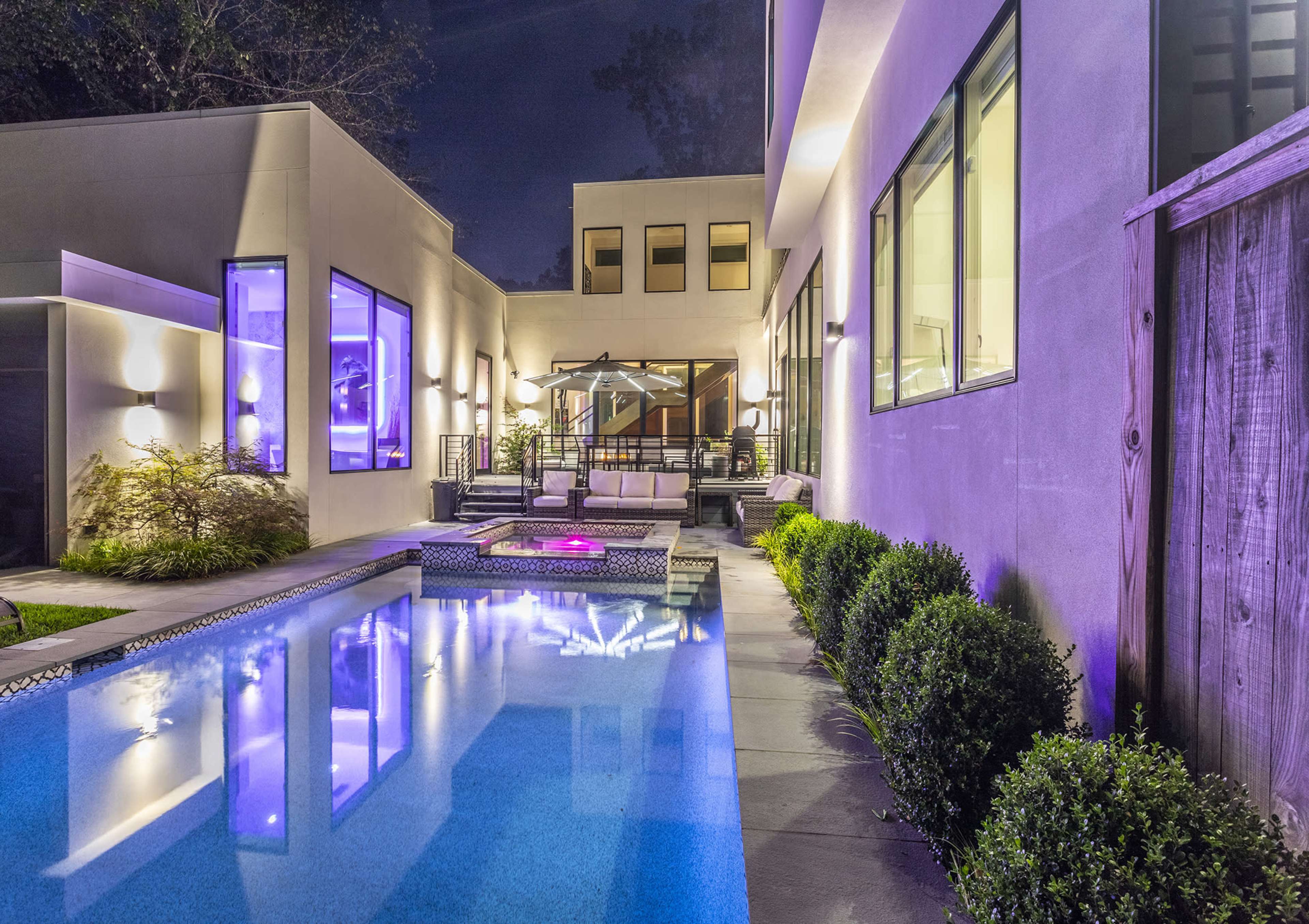 A modern home features a swimming pool illuminated with purple lights, surrounded by landscaped greenery and large windows at night.