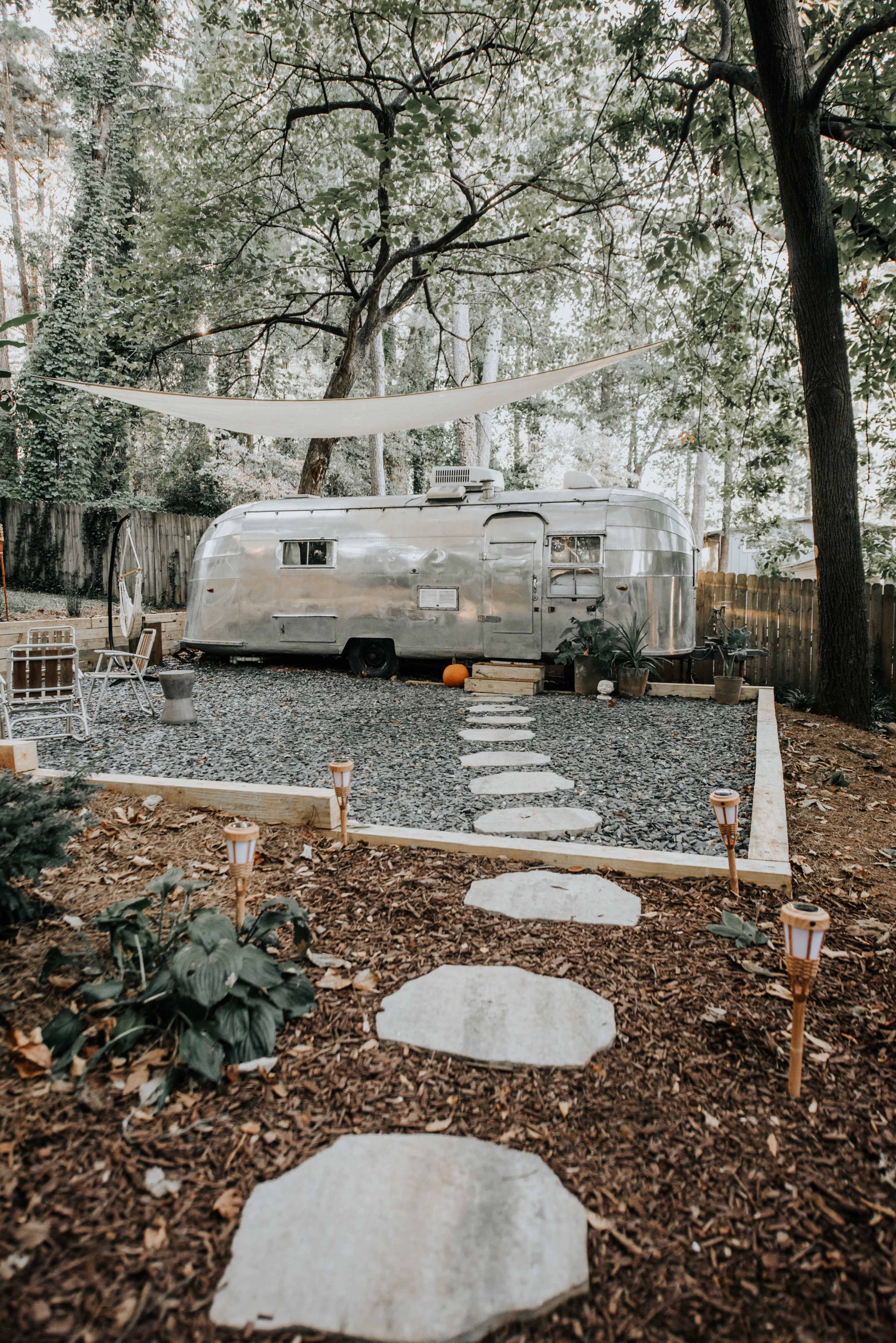 A silver Airstream trailer is parked in a gravel area surrounded by trees, with a stone path leading to it and outdoor seating nearby.