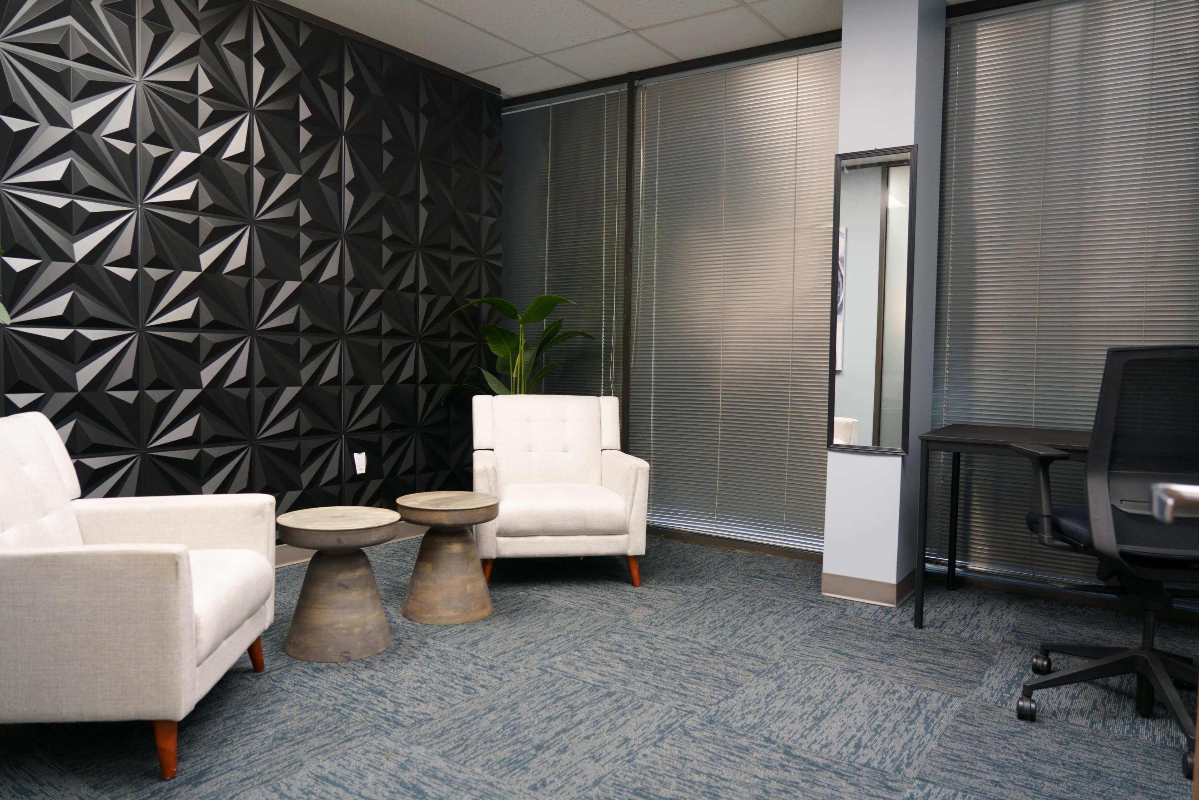 The image shows a modern office space featuring two white upholstered chairs, two round metal side tables, a small indoor plant, and a desk with a black chair against a textured black wall and partially windowed walls.