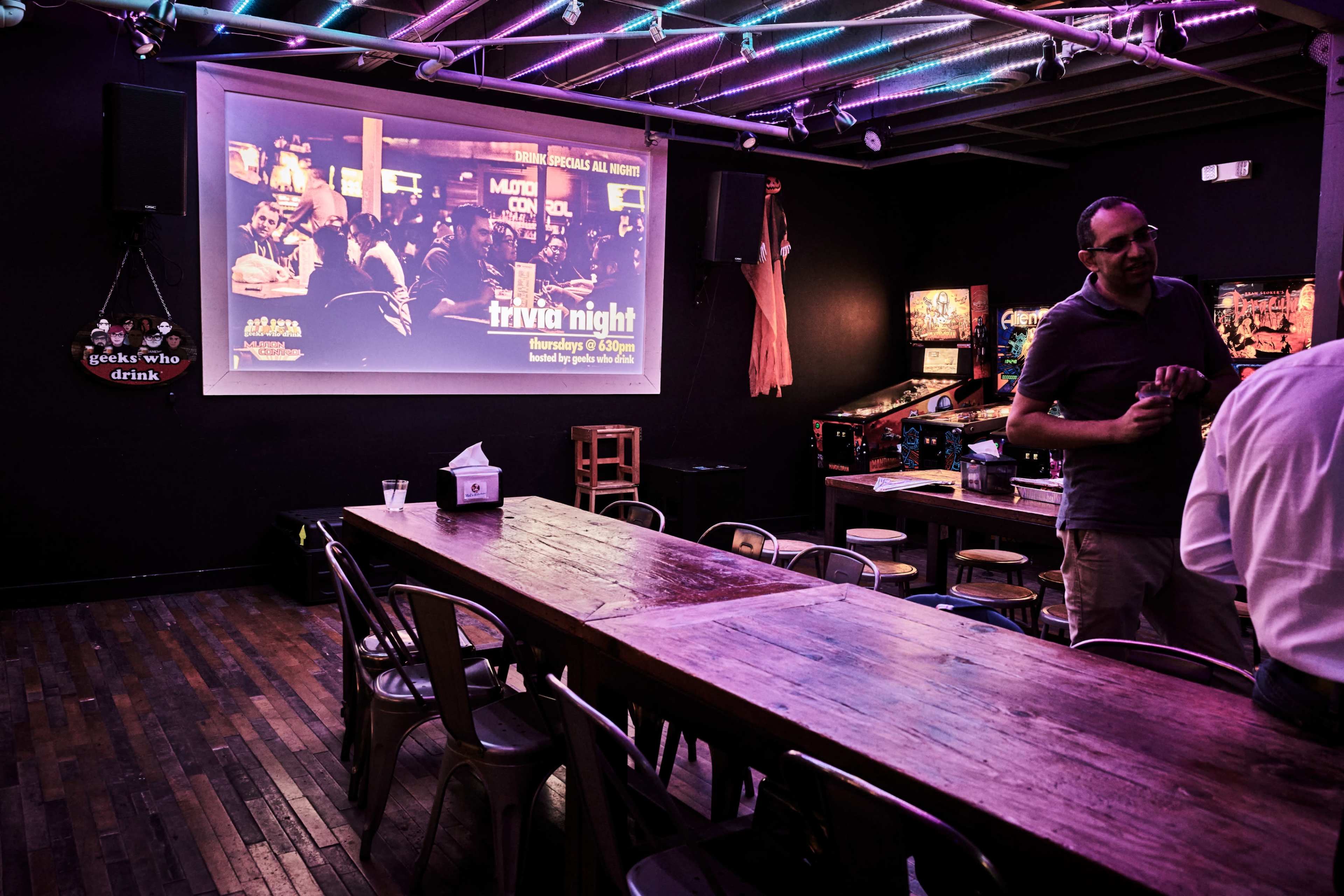 A dimly lit bar with a long wooden table and a projector screen displaying a trivia night advertisement on the wall.