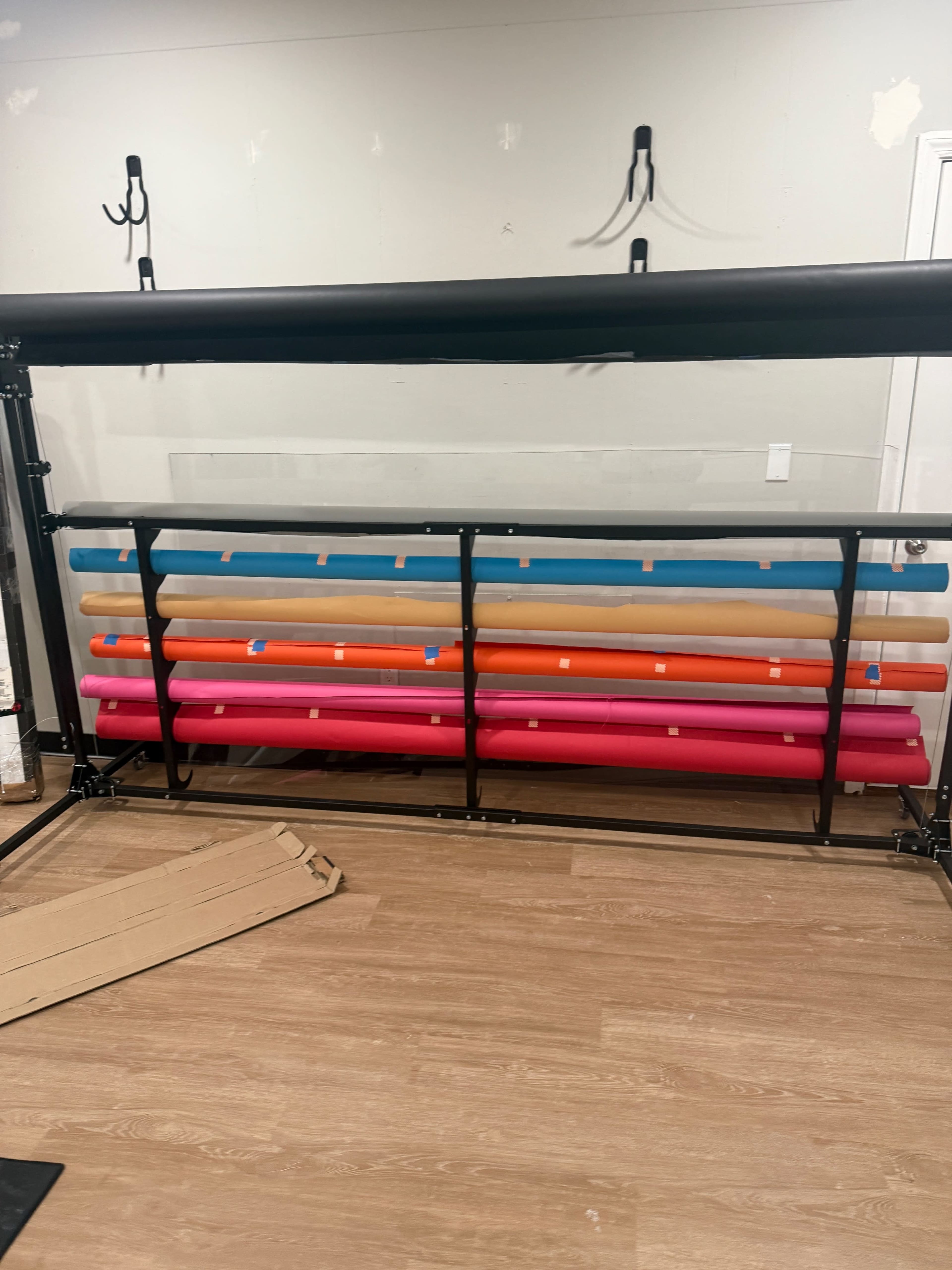 The image shows a storage rack holding various rolls of colored vinyl in a room with wooden flooring and some wall hooks.
