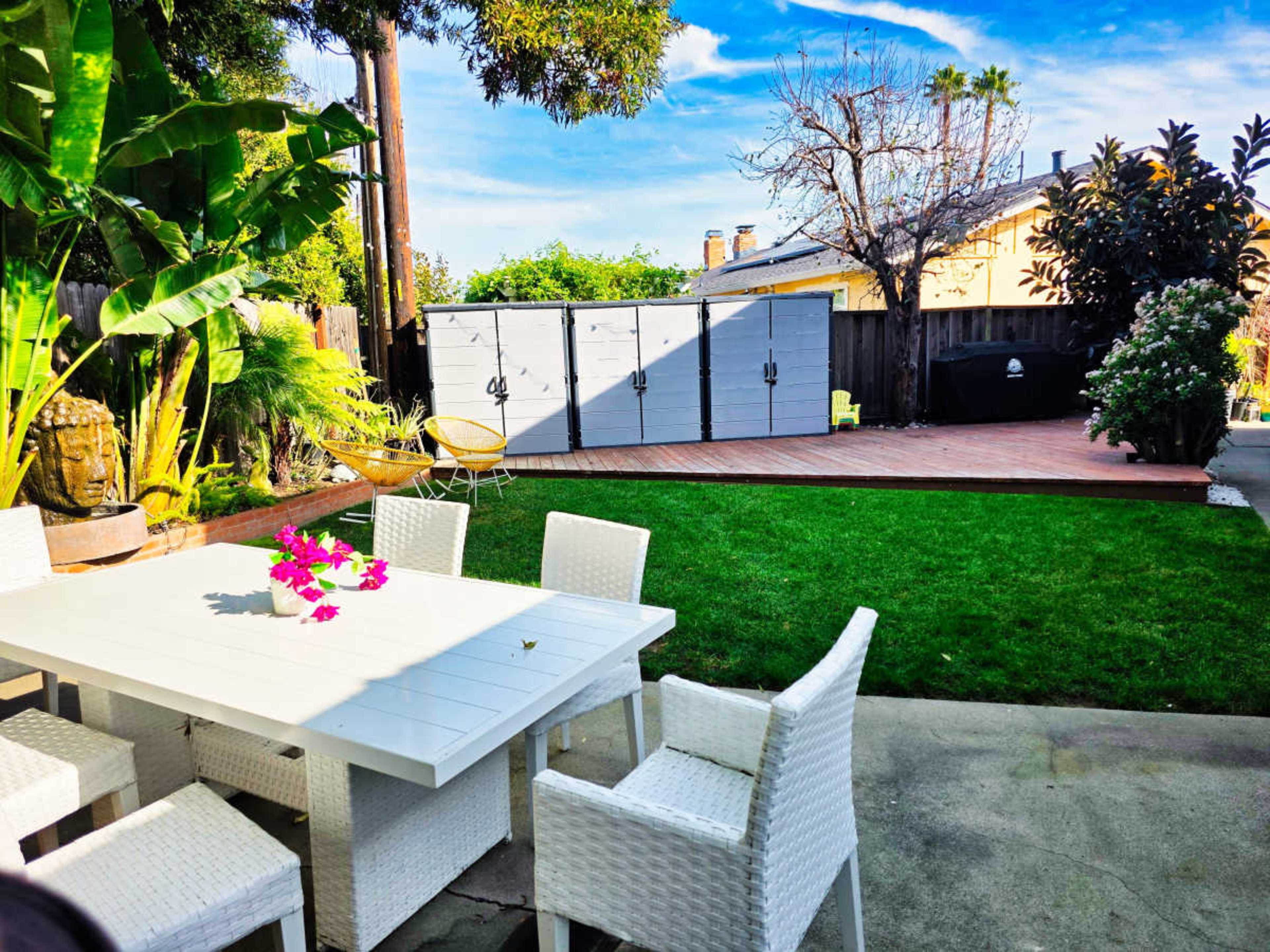 The image shows a backyard with a white dining table and chairs, green grass, and storage sheds in the background under a clear blue sky.